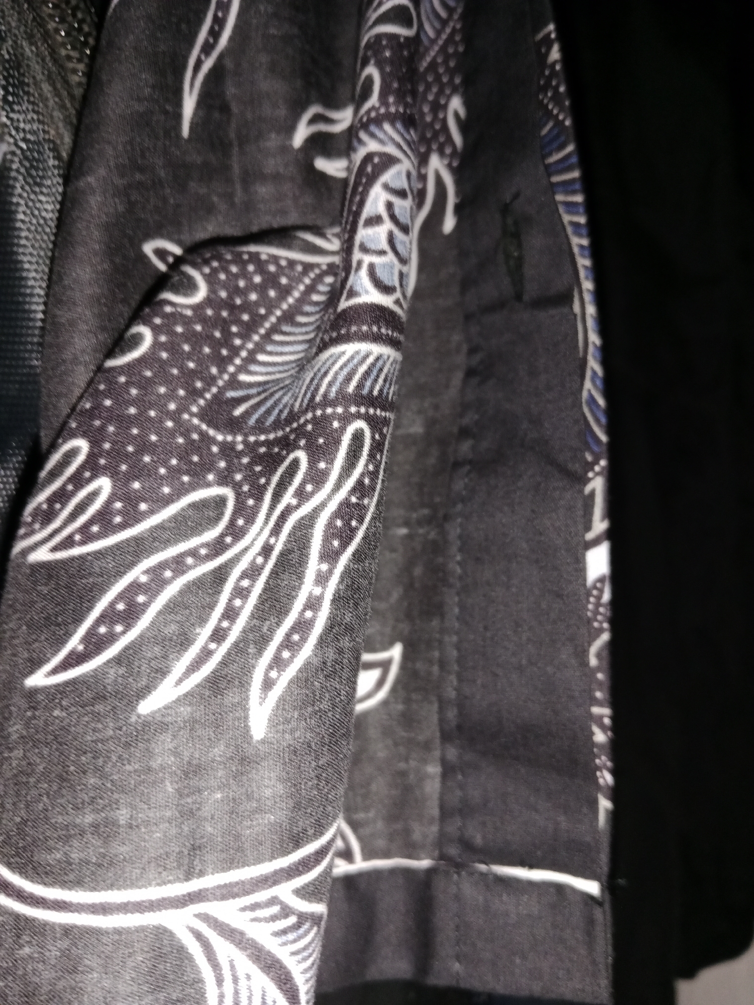 The image shows a close-up of a black fabric with a white and blue batik pattern. The fabric is draped and folded, creating shadows and highlights that emphasize the texture of the material. The batik design features swirling lines and small dots, creating a visually intricate pattern. The setting appears to be indoors, possibly a clothing store or a home in Ringinrejo, Indonesia, given the batik pattern is a traditional Indonesian art form. No people or specific actions are visible.