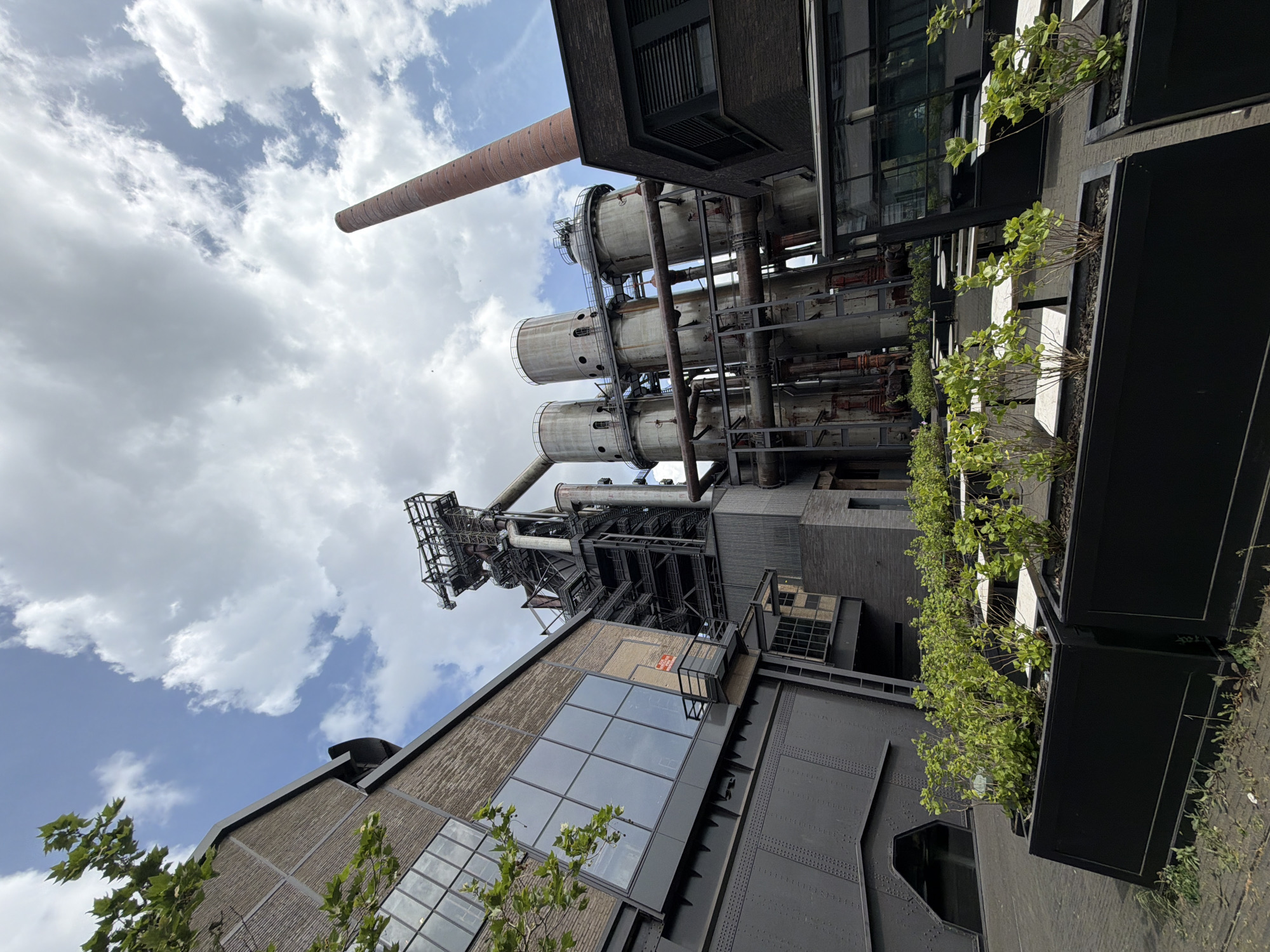 The image depicts an industrial complex in Esch-sur-Alzette, Luxembourg, set against a cloudy sky. A tall, brick chimney rises on the left, adjacent to a series of vertical cylindrical structures connected by a framework of pipes and metal beams. A building constructed with dark metal and brick, featuring a grid of windows, is visible below the industrial structures. Planter boxes filled with greenery line the edge of a walkway.