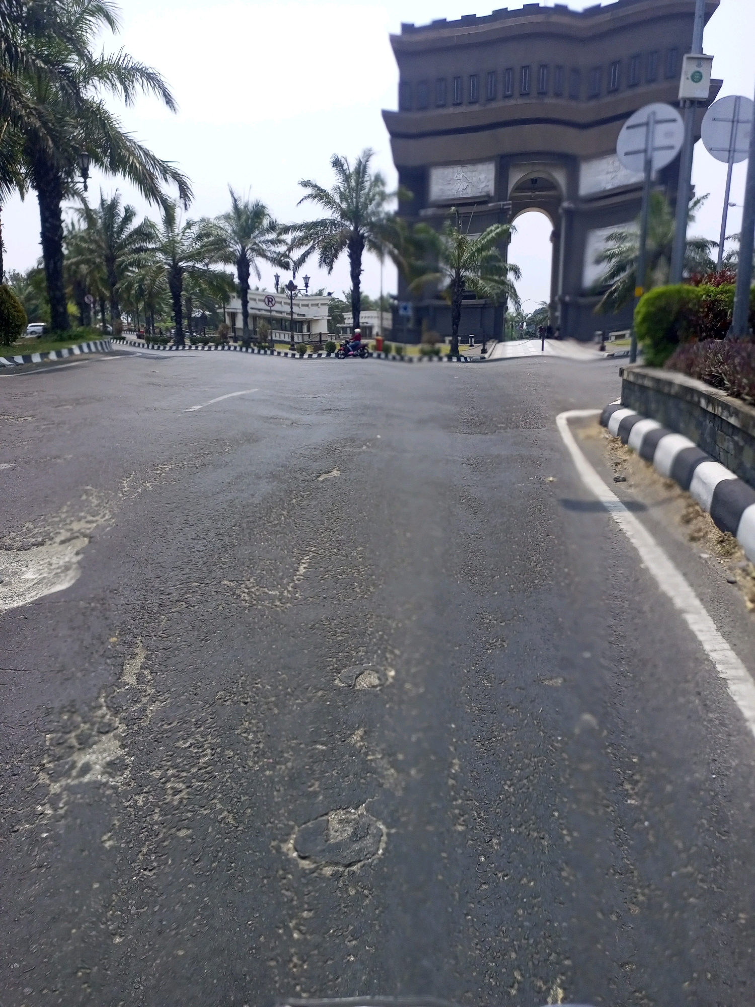 The image depicts a street scene in the city of Ngasem, Indonesia, characterized by an archway structure in the background and palm trees lining the road. In the foreground, the asphalt road surface exhibits signs of wear and tear, with visible patches and cracks. A white line marks the center of the road. To the right, a paved sidewalk is bordered by a decorative wall with black and white striped curbing. A person on a pink motorcycle is visible on the right side of the road, moving away from the camera. In the distance, the large, ornate archway serves as a prominent landmark. The sky is overcast, suggesting a cloudy day.