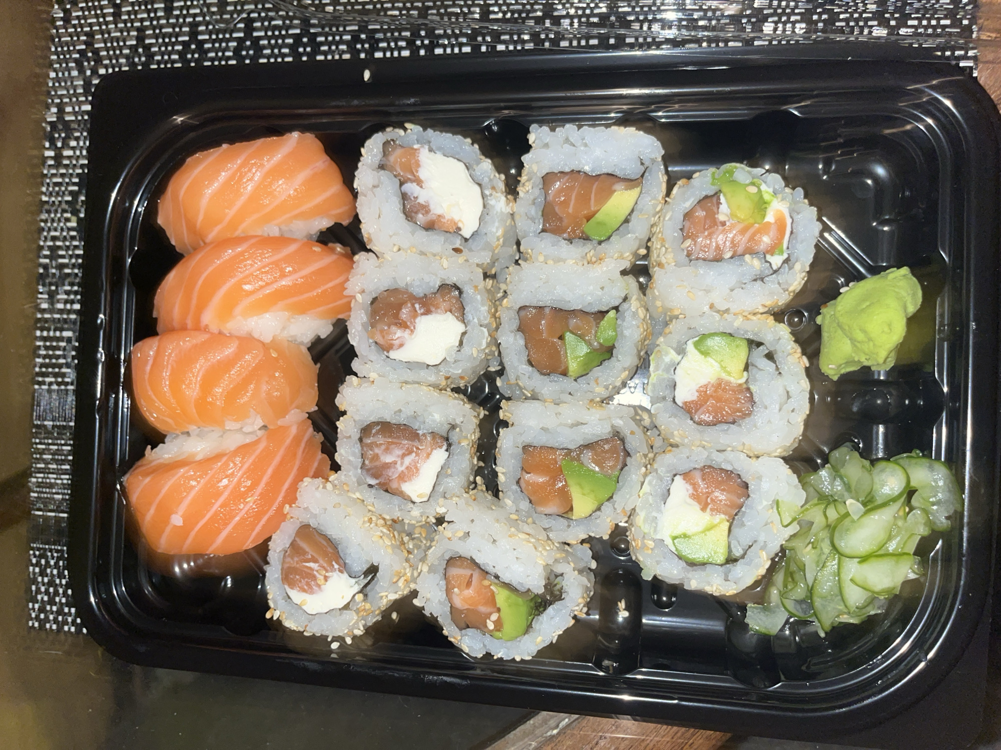 A black plastic container holds an assortment of sushi. On the left are four pieces of salmon nigiri. Adjacent to the nigiri are six California rolls filled with salmon and cream cheese. To the right of those rolls are six California rolls filled with salmon and avocado. A scoop of wasabi and a mound of marinated cucumbers are in the upper and lower right corners, respectively. The tray rests on a textured, dark-colored surface, possibly a placemat or table. The location is not discernible from the image.