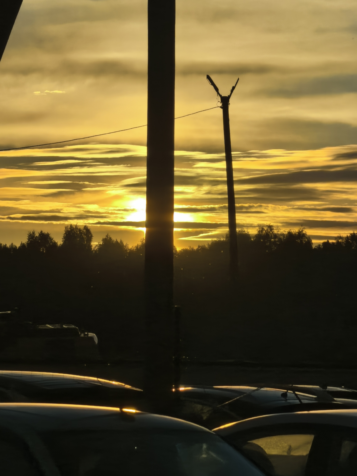The photograph captures a striking sunset scene silhouetted against a dramatic sky. The sun, a brilliant orb of yellow, is low on the horizon, casting long rays through horizontal cloud formations that streak across the sky in shades of gold and yellow. In the foreground, two large, dark poles stand prominently, one to the left and another slightly to the right and further back, with a detached Y-shaped fixture atop the latter, resembling a street lamp or utility pole. Between the poles and the horizon, a dark, dense line of trees forms a silhouetted treeline. Below the treeline, the foreground is filled with the dark silhouettes of parked cars, their shapes defined by the reflected glow of the sunset on their surfaces. The overall impression is one of a dramatic natural event juxtaposed with everyday urban infrastructure.