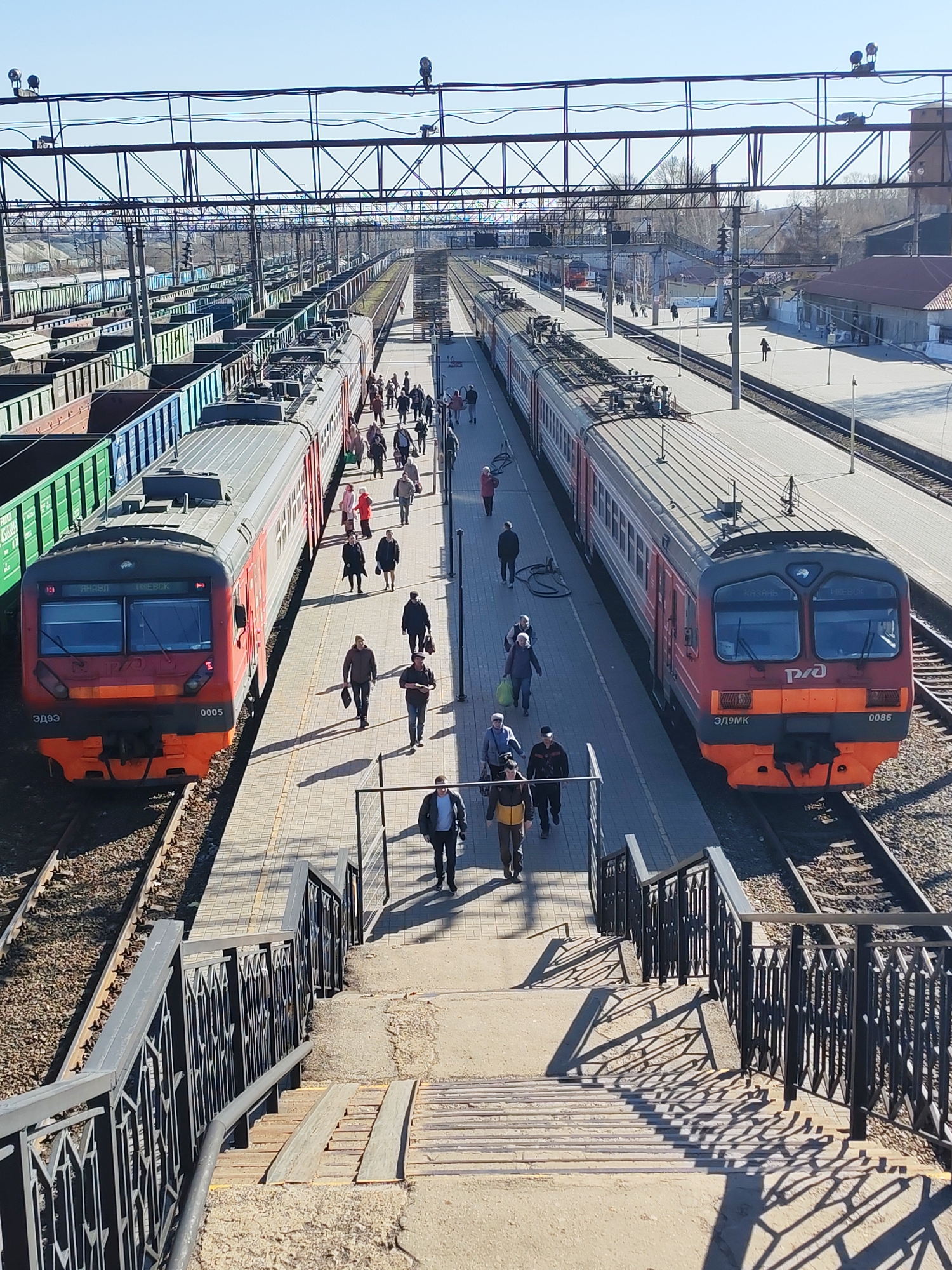 An elevated, high-angle view captures a busy train station in Izhevsk, Russia, on a bright, sunny day. The scene features multiple railway tracks, platforms, and trains under a clear blue sky, with the sun casting long, distinct shadows, suggesting morning or late afternoon.
In the foreground, a set of concrete stairs with ornate metal railings descends towards the platforms. Directly below, two passenger electric multiple unit (EMU) trains are stopped at platforms, facing away from the viewer.
The train on the left platform is an ЭД9Э (ED9E) series, marked with the number "0005" on its front. Its livery is red and orange with a gray roof and white window bands. Destination signs above its windows show "ЯНАУЛ" (Yanaul) and "ИЖЕВСК" (Izhevsk). Alongside this platform, to the far left, a long line of freight wagons, primarily open-top hoppers in green and brown, are parked on an adjacent track, with some faint "РУСАЛ" text visible on one.
The train on the right platform is an ЭД9МК (ED9MK) series, bearing the number "0086". It shares a similar red and orange livery. Its destination signs indicate "КАЗАНЬ" (Kazan) and "ИЖЕВСК" (Izhevsk).
Both platforms are populated by numerous people walking, likely commuting or traveling. They appear to be boarding, alighting, or waiting for trains. The overall atmosphere is one of active transit. Overhead, a complex network of catenary wires and supporting gantries spans across the tracks. In the background, beyond the platforms, more tracks, buildings, and distant trees are visible, along with another train further down the line.