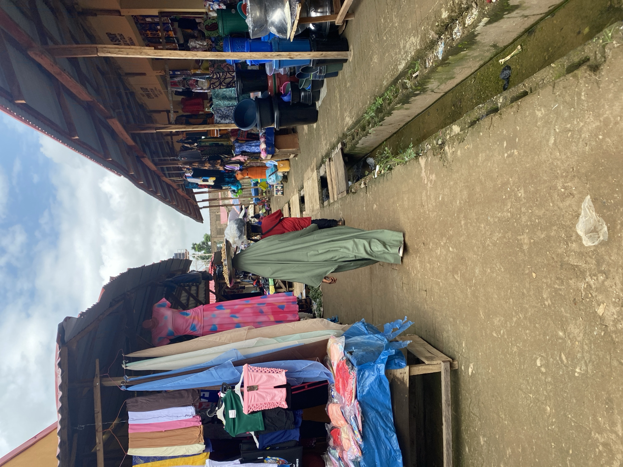This is an outdoor market scene in Kafanchan B, Nigeria. The scene depicts a narrow walkway lined with stalls selling clothing and various household items. The stalls have corrugated metal roofs supported by wooden beams. On the left, clothing items such as dresses, shirts, and shorts are displayed on hangers and on a table covered with a blue plastic sheet. On the right, numerous plastic buckets and containers are stacked. In the center of the walkway, a person wearing a green headscarf and flowing garment walks away from the camera, carrying a flat basket on their head. Another person in a red shirt is visible behind them. A concrete drainage ditch runs along the right side of the walkway, containing murky water and some debris. The ground is paved with concrete. The sky is partly cloudy.