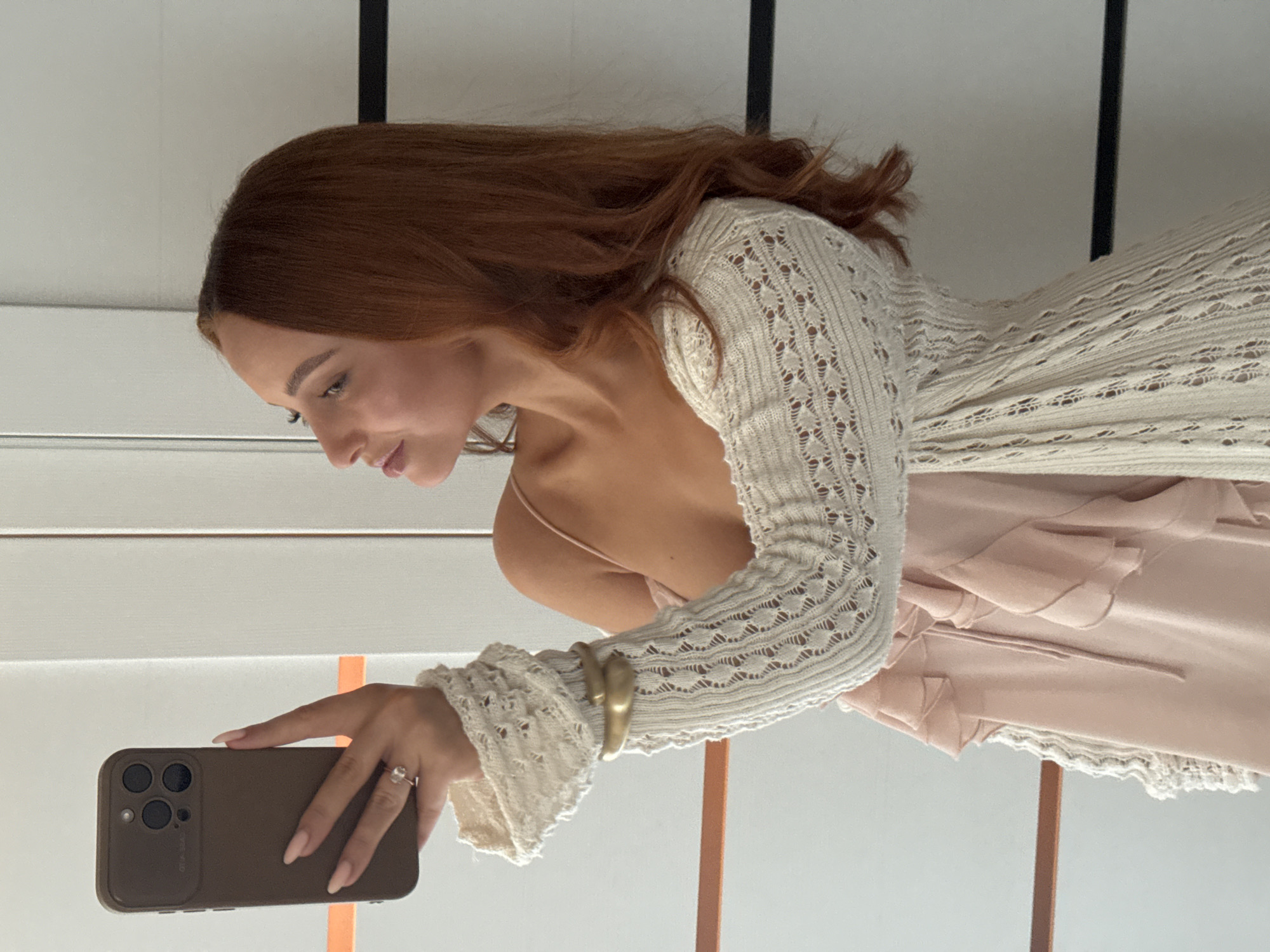 A woman with auburn hair is taking a selfie in an indoor setting. She is wearing a light pink dress with ruffles and a cream-colored crocheted cardigan with bell sleeves. She has a gold cuff bracelet on her left arm and a ring on her left ring finger. Her right hand is holding a brown smartphone, pointed towards a mirror. The background appears to be a wall with vertical white panels and some horizontal orange and black accents. There is no indication of the city of Chaikovsky, Russia, in the image.