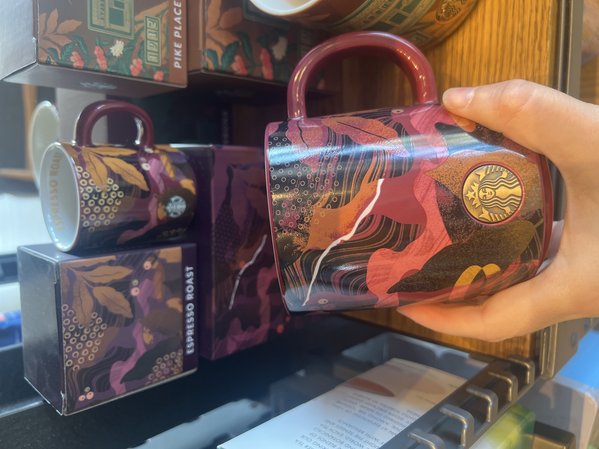 A hand holds a Starbucks mug decorated with a colorful botanical design featuring pink and orange leaves against a dark background. The mug also has a gold Starbucks logo. In the background, there are two Starbucks coffee boxes, one labeled "ESPRESSO ROAST" and another partially visible box with "PIKE PLACE" text. The scene appears to be inside a Starbucks store, likely in the city of Ko Samui District, Thailand, given the context.