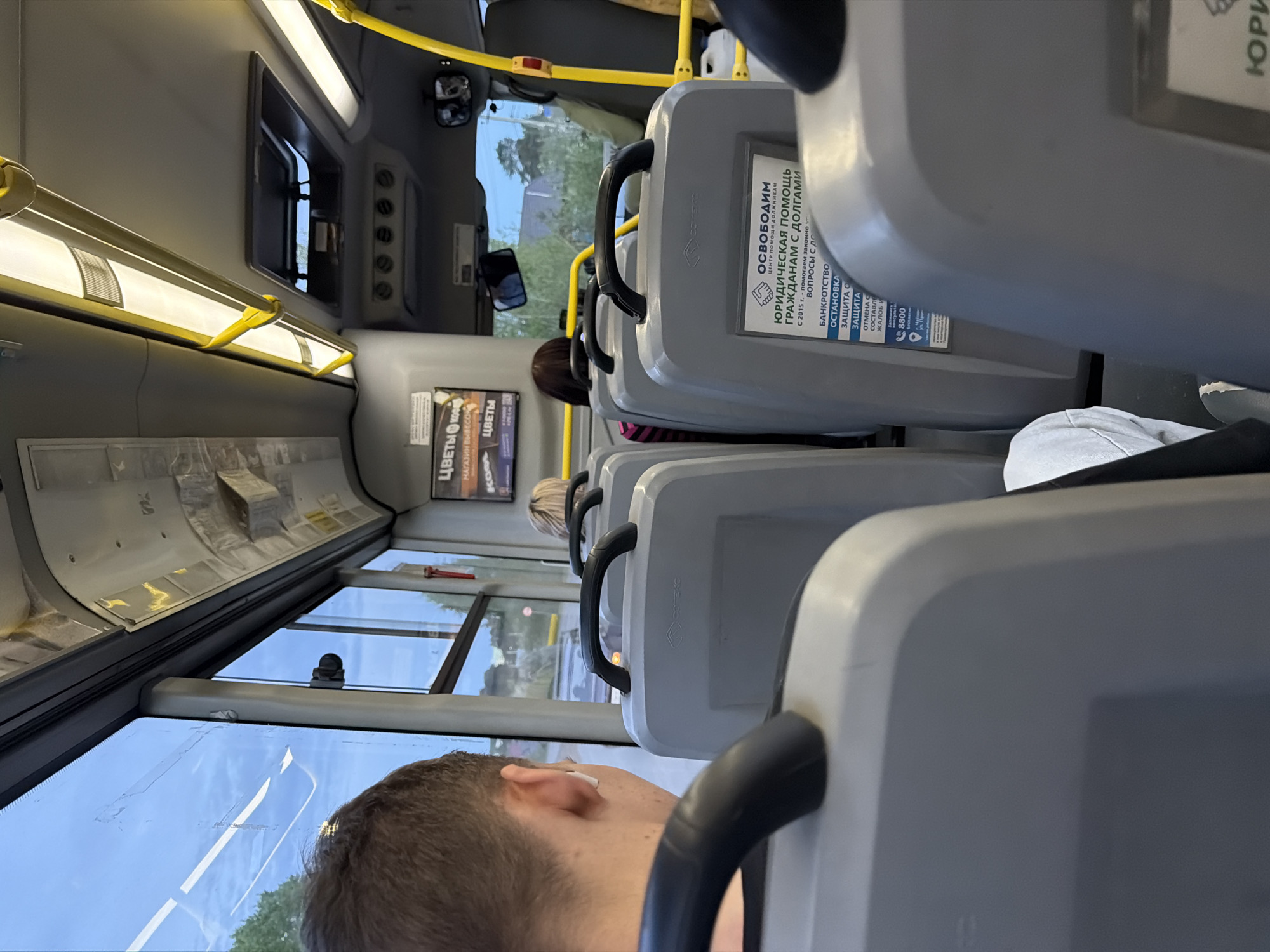 The image, taken inside a bus in what appears to be Chaikovsky, Russia, shows a row of grey bus seats. A man's head is visible in the foreground. Across the aisle, advertisements are pasted to the back of the seats. The text "ЦВЕТЫ" and "КОФЕ" are visible on one advertisement. Another advertisement provides assistance for citizens with debts and includes services such as bankruptcy and defense. Outside the bus windows, trees and buildings are visible. A yellow handrail runs along the ceiling of the bus.