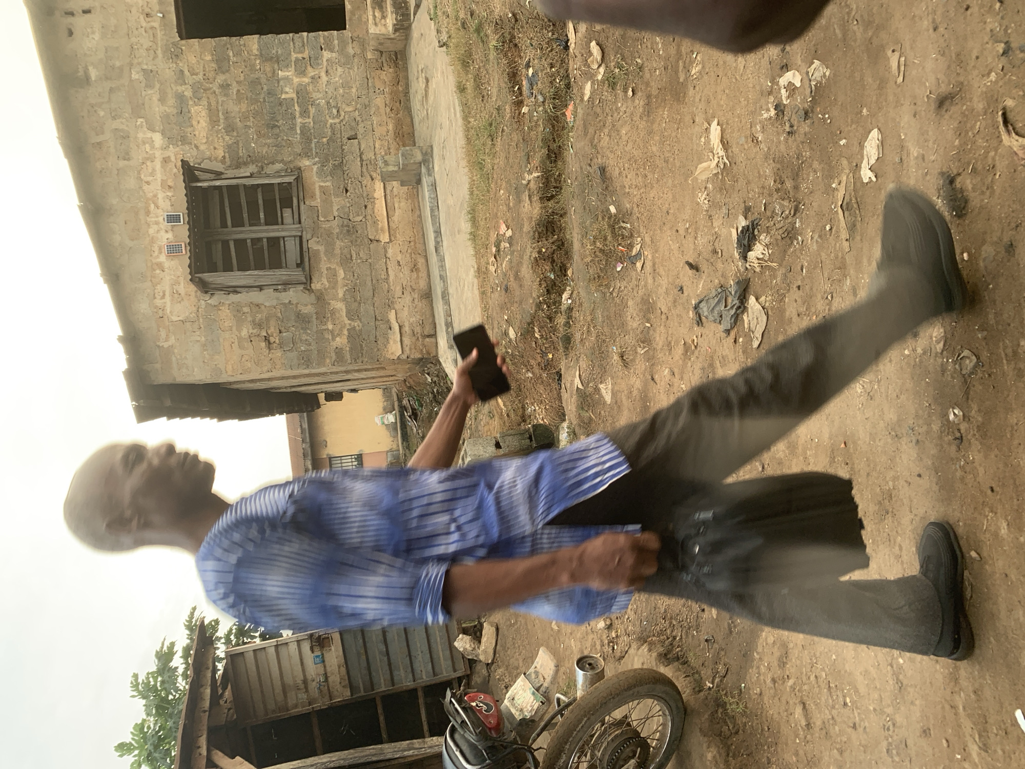 A man walks down a dirt path in Shagamu, Nigeria, carrying a black plastic bag in his left hand and a smartphone in his right. He is wearing a blue and white striped shirt and dark pants. A motorcycle is parked to his left, and a brick building with solar panels on the roof is visible in the background. The ground is littered with trash. The scene appears to be in an urban or semi-urban area.