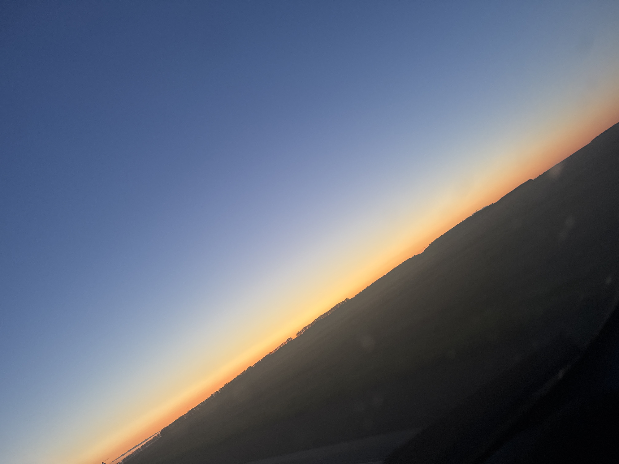 The image depicts a diagonal view of a sunset over a landscape, taken from an elevated perspective, possibly an airplane. The sky transitions from a deep blue at the top right to a band of orange and yellow near the horizon line. The horizon itself is marked by a dark, silhouetted landscape that appears to be a treeline, suggesting a rural or semi-rural area. The foreground on the left is dominated by a dark, shadowed area, which could be part of the aircraft or a structure. The scene is devoid of any people or identifiable structures within the visible landscape, creating a sense of natural beauty and isolation. The location is identified as the city of Ploskaya, Russia, during twilight.