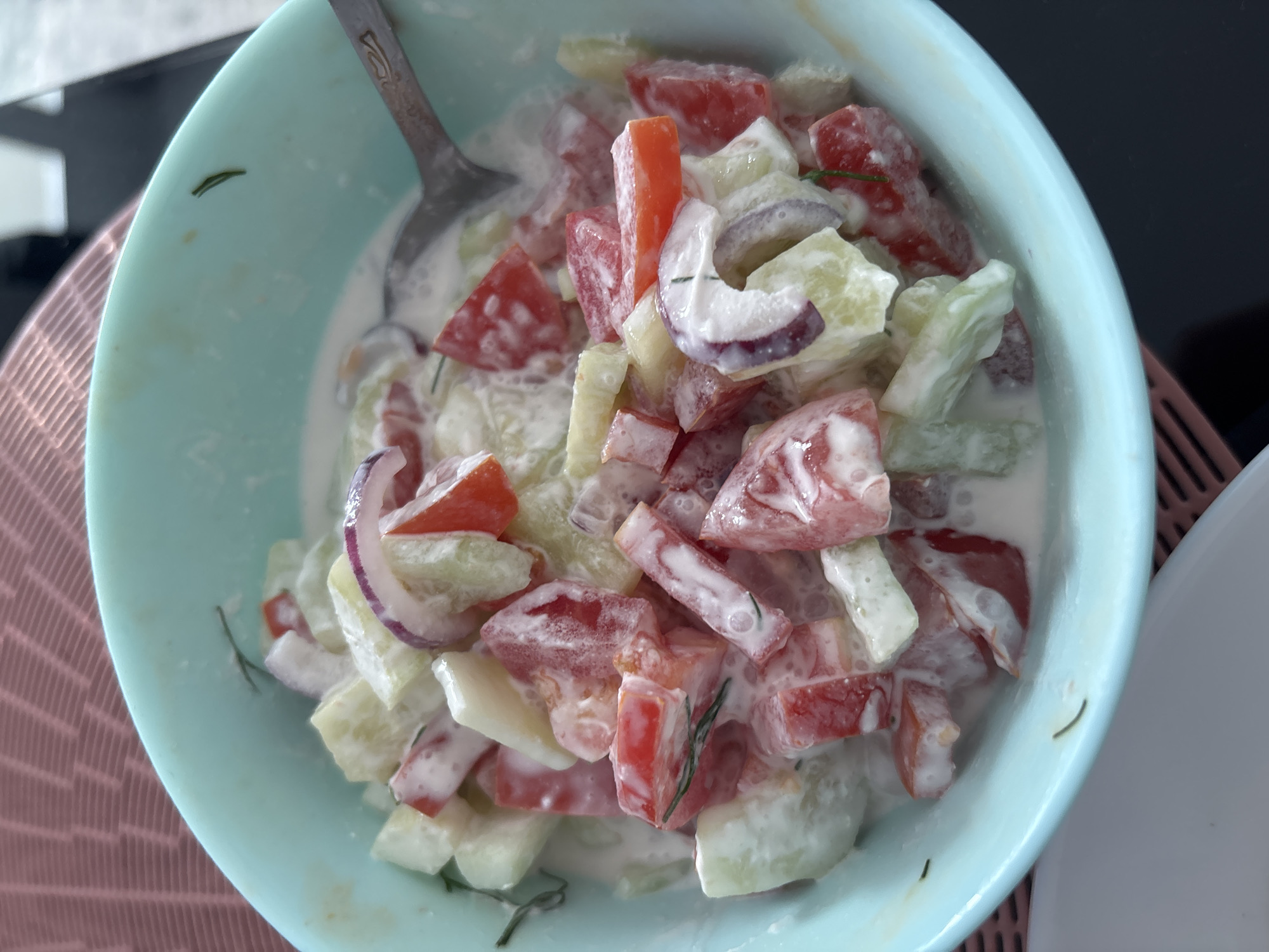 A close-up, overhead shot shows a light blue bowl filled with a chopped salad. The salad consists of diced tomatoes, cucumbers, red onion rings, and possibly dill, all coated in a creamy white dressing. A silver spoon rests in the bowl on the right side of the frame. The bowl is placed on a pink textured placemat, which is on a dark surface. The scene is indoors, likely in a home kitchen in Minsk, Belarus.