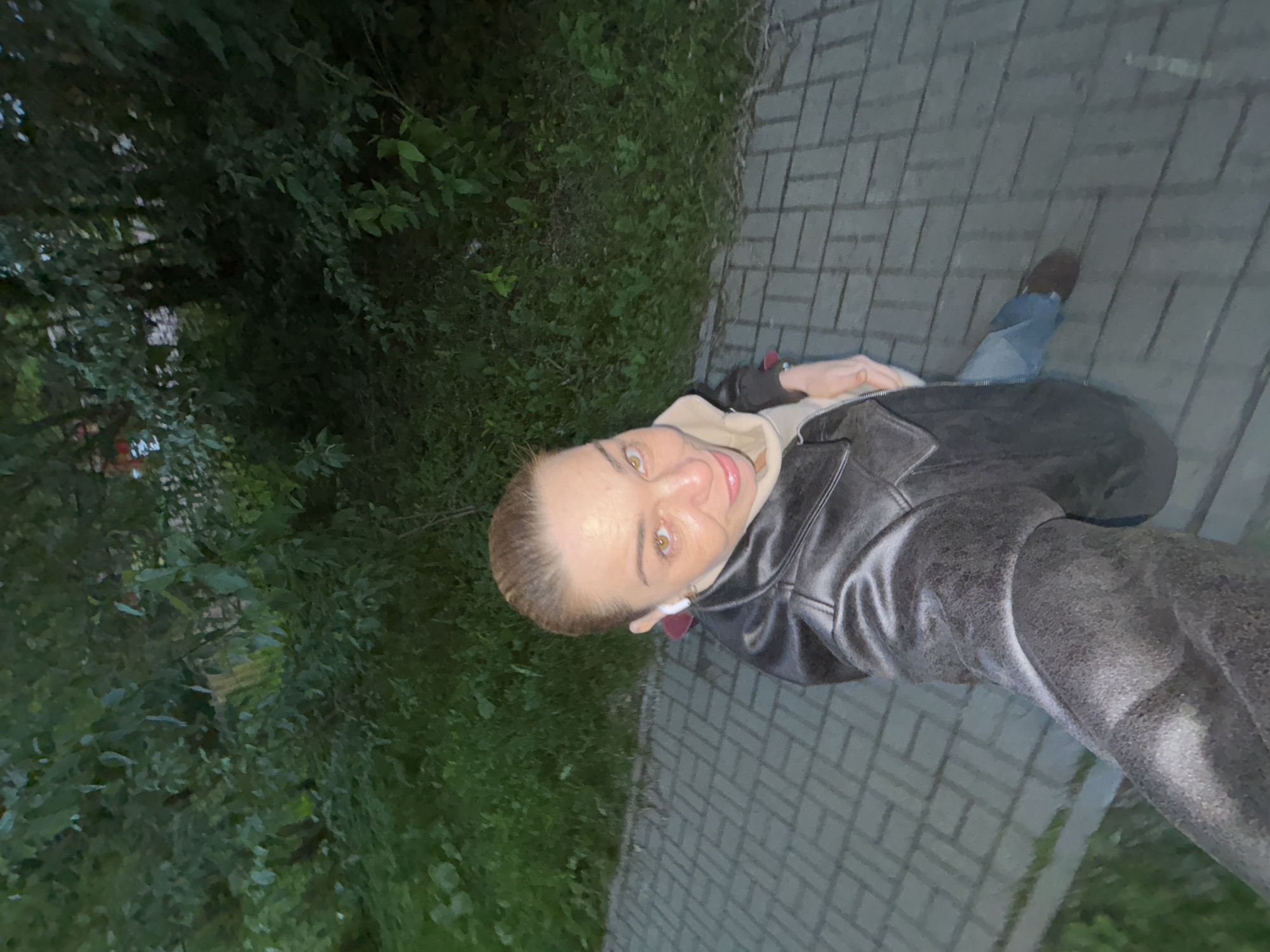 A woman is taking a selfie on a brick pathway next to a grassy area with dense trees. She is wearing a black leather jacket, a light beige hoodie, and blue jeans. She has light skin, brown eyes, and her hair is pulled back. She is also wearing white wireless earbuds. The scene appears to be outdoors in a park or residential area, possibly in the evening or early morning given the low light.