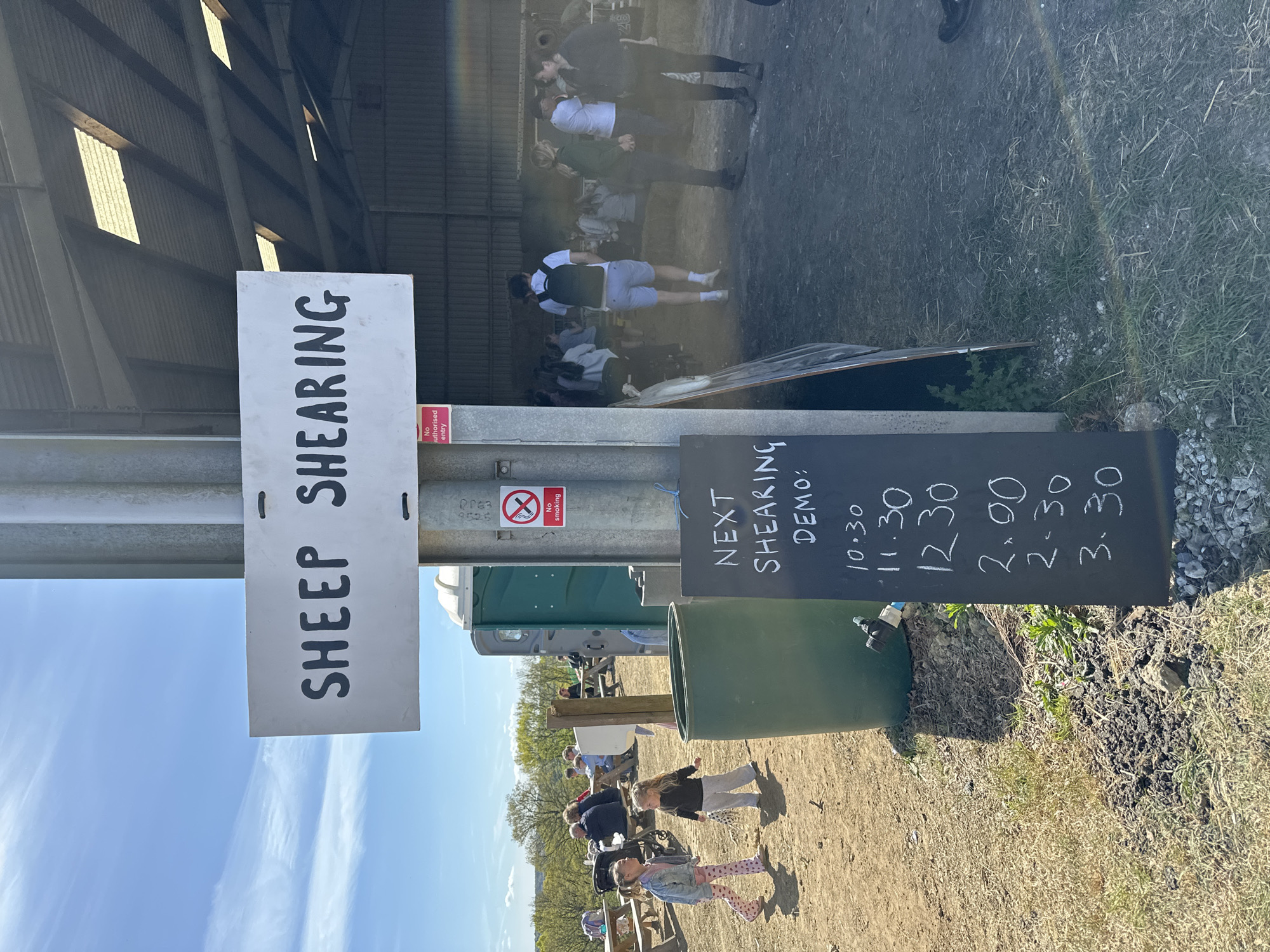 The image shows an outdoor scene with a sign that reads "SHEEP SHEARING" vertically. Adjacent to it is a chalkboard indicating "NEXT SHEARING DEMO:" followed by times: 10.30, 11.30, 12.30, 2.00, 2.30, and 3.30. A metal post supports both signs. The background reveals a partially open building with people inside. In the foreground, there's a green water tank, two young girls in casual clothes, and picnic benches occupied by people. There is a portable toilet and 'No Smoking' sign are also visible. The environment includes dirt, gravel, and grass. The location is likely Sevenoaks, United Kingdom, based on identifying marks on the signs.