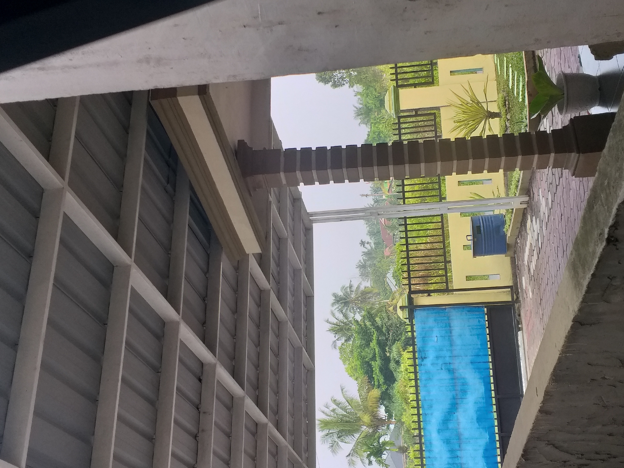 The image depicts the exterior of a property in Air Jamban, Indonesia. A corrugated metal roof supported by white beams is visible in the foreground, casting shadows. A substantial brown, octagonal pillar stands to the right, partially obscuring the view. Beyond this structure, a black metal fence with yellow posts and a large blue gate with a textured surface are prominent. In the background, lush green trees, including palm trees, create a dense foliage. Several houses with tiled roofs can be seen amidst the greenery. The ground is paved with interlocking stones, and a small garden area with stepping stones and plants, including a potted plant, is situated near the fence. A blue plastic barrel is also present in the garden. The overall scene suggests a residential area with tropical vegetation under an overcast sky.