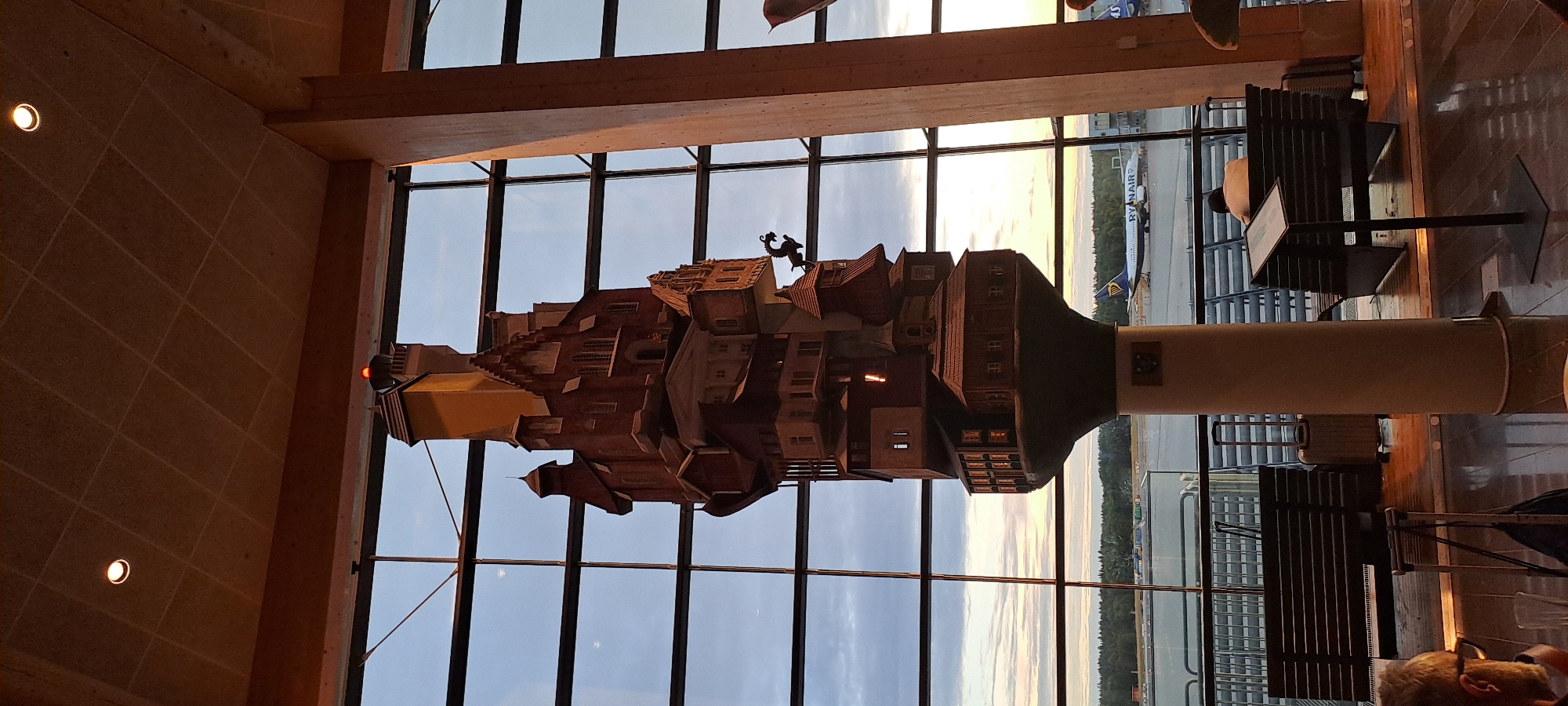 This image captures a view from inside Stockholm-Arlanda Airport, likely within a lounge area, overlooking the tarmac. A prominent, multi-layered sculpture resembling a whimsical, stacked village or castle dominates the center foreground, silhouetted against a large window. The sculpture is made of wood and features numerous windows, roofs, and architectural details. A red light is visible at the very top of the sculpture.
Through the expansive window, the airport tarmac is visible, with a Ryanair airplane taxiing or parked in the distance. The sky is a muted twilight, with hints of sunset colors. In the foreground, seating benches with metal slats are visible, along with a few pieces of luggage, including a silver suitcase and a black carry-on bag. A man wearing glasses is partially visible on the left, and another person is seated on a bench on the right, facing away from the camera. The overall environment suggests an indoor waiting area at an airport, with natural light streaming in from the large windows.