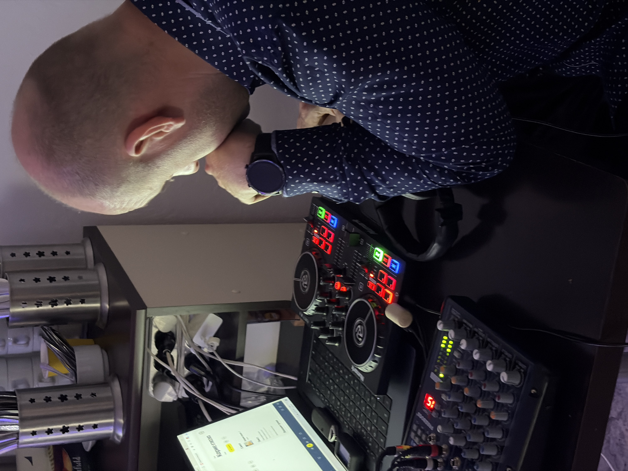 A balding man with a dark blue patterned shirt and a wristwatch is looking at a desk with a computer, a DJ controller, a mixer, and headphones. The desk is adjacent to a countertop with kitchen utensil holders. The location cannot be confirmed as Votkinsk, Russia from the image.