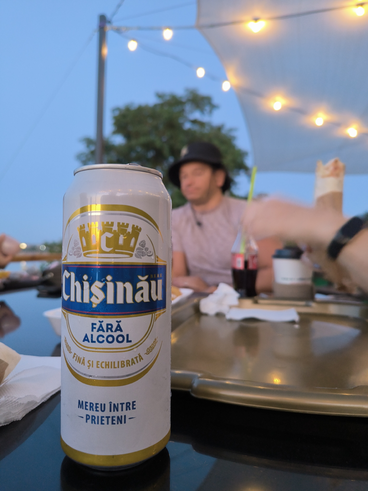 A Chisinau brand non-alcoholic beverage can is centered in the foreground, positioned on a reflective dark surface, possibly a table. The can features a white and gold label with blue accents. The prominent text on the label reads "Chisinau," followed by "FĂRĂ ALCOOL" (without alcohol) and "FINĂ ȘI ECHILIBRATĂ" (fine and balanced). Below this, it states "MEREU ÎNTRE - PRIETENI -" (always among friends). The can appears slightly dewy.
In the middle ground, a person wearing a dark hat is seated, with their face slightly out of focus. To their right, a bottle of Coca-Cola with a green straw is visible, along with a white paper cup and what appears to be food, possibly a wrapped sandwich or kebab. Another person's hand, wearing a black smartwatch, is reaching towards the right side of the frame.
The background is blurred, showing an outdoor setting with string lights illuminating the scene, consistent with an evening ambiance. Trees and a white canopy are also discernible. The overall impression is a casual outdoor gathering.
While the image depicts a Chisinau brand beverage, there is no direct indication or visual information within the image to confirm the specific location as Ustia, Moldova.