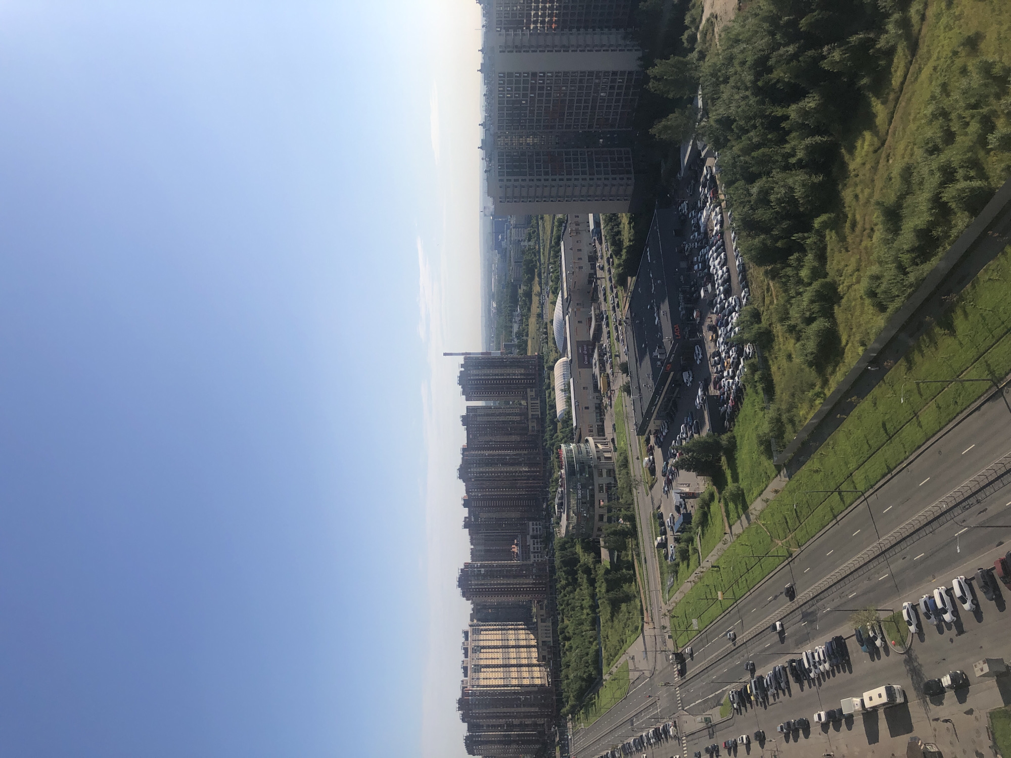 The image depicts a sunny day in an urban environment with a clear blue sky. In the foreground, a multi-lane road runs diagonally from the bottom left to the middle of the frame, with numerous cars parked along its right side and a few vehicles in motion. Adjacent to the road on the right is a strip of green grass with streetlights. Further into the scene, to the right of the road, is a large parking lot filled with cars, situated in front of a dark-colored building with the word "LADA" visible, suggesting a car dealership. Beyond this, a dense area of green trees and shrubs occupies the middle ground, with a circular building with glass walls and text reading "TOKYO-CITY" visible behind it. In the background, a dense cluster of tall, modern residential buildings dominates the landscape, extending across the horizon. To the far left of the background, a tall, industrial-looking structure with a red top is visible. The overall scene suggests a developed suburban or exurban area with a mix of residential, commercial, and green spaces.