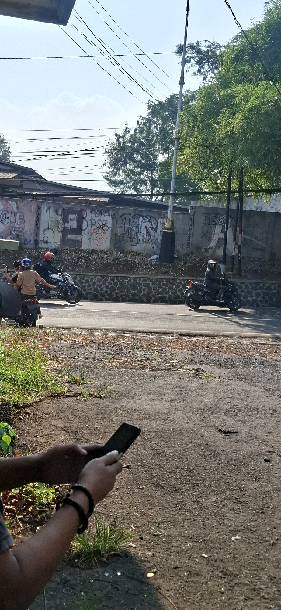 The image depicts a street scene in Cilaku, Indonesia, on a clear day. In the foreground, a person's hands are visible, holding a black smartphone. They are wearing a dark bracelet. The background shows a paved road where several motorcycles are traveling.
On the left side of the road, two people are riding a motorcycle. The rider wears a helmet, and the passenger, seen from the back, is wearing a light brown shirt and a blue helmet, and appears to be taking a photo with their phone. To the right of this motorcycle, another person is riding a motorcycle, also wearing a helmet and a dark jacket.
Behind the road, a stone wall runs horizontally, topped by a concrete wall covered in graffiti. The graffiti includes numbers and words that are partially obscured and illegible. A tall, slender pole with electrical wires running across it is visible against the sky. Lush green trees and vegetation are present behind the wall, some of which appear to be bamboo. The overall impression is a snapshot of everyday life on a street in Cilaku.