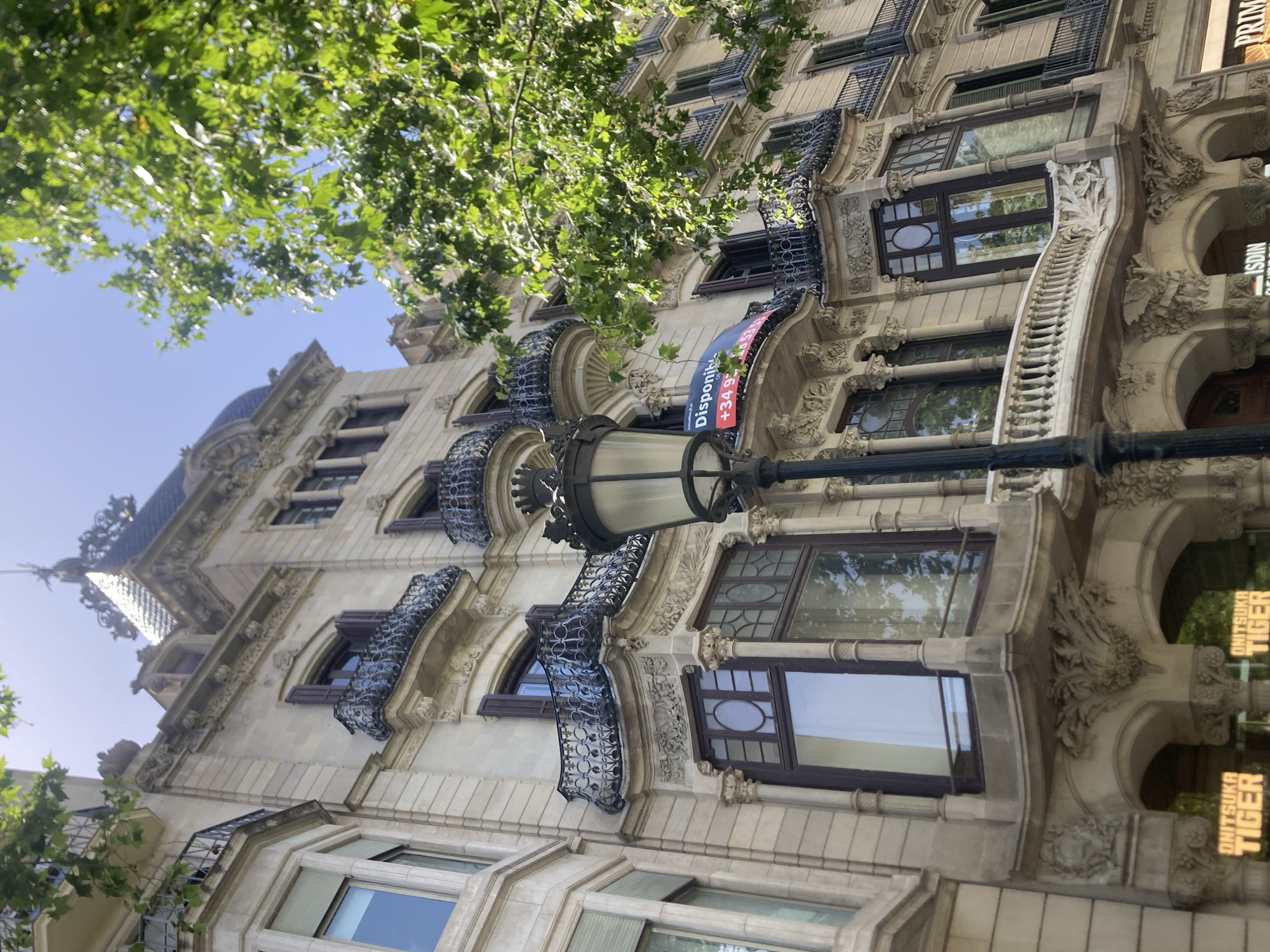 The image shows a close-up, low-angle view of an ornate building facade, likely in Barcelona, Spain, given the text visible on a banner and on a storefront. The architecture features intricate stone carvings, decorative balconies with wrought-iron railings, and arched windows. Lush green leaves from a tree overhang the top of the building, suggesting it's daytime and the weather is pleasant.
The building's facade is made of light-colored stone, with dark-framed windows that reflect the surrounding greenery and sky. Several balconies jut out from the building, each adorned with decorative metalwork. The building's design appears to be in a historic style, possibly Art Nouveau or a similar ornate architectural movement.
Visible text includes a blue and red banner on the building that reads "Disponil" followed by what appears to be a phone number, "+34 9...". Further down, on a storefront visible through an arched opening, the text "ONITSUKA TIGER" is clearly displayed in yellow, indicating a retail establishment.
A streetlamp with a classic design is positioned in the foreground, partially obscuring the view of the building. The overall impression is one of an architecturally significant building in a vibrant urban setting on a sunny day. No people are visible in the image.