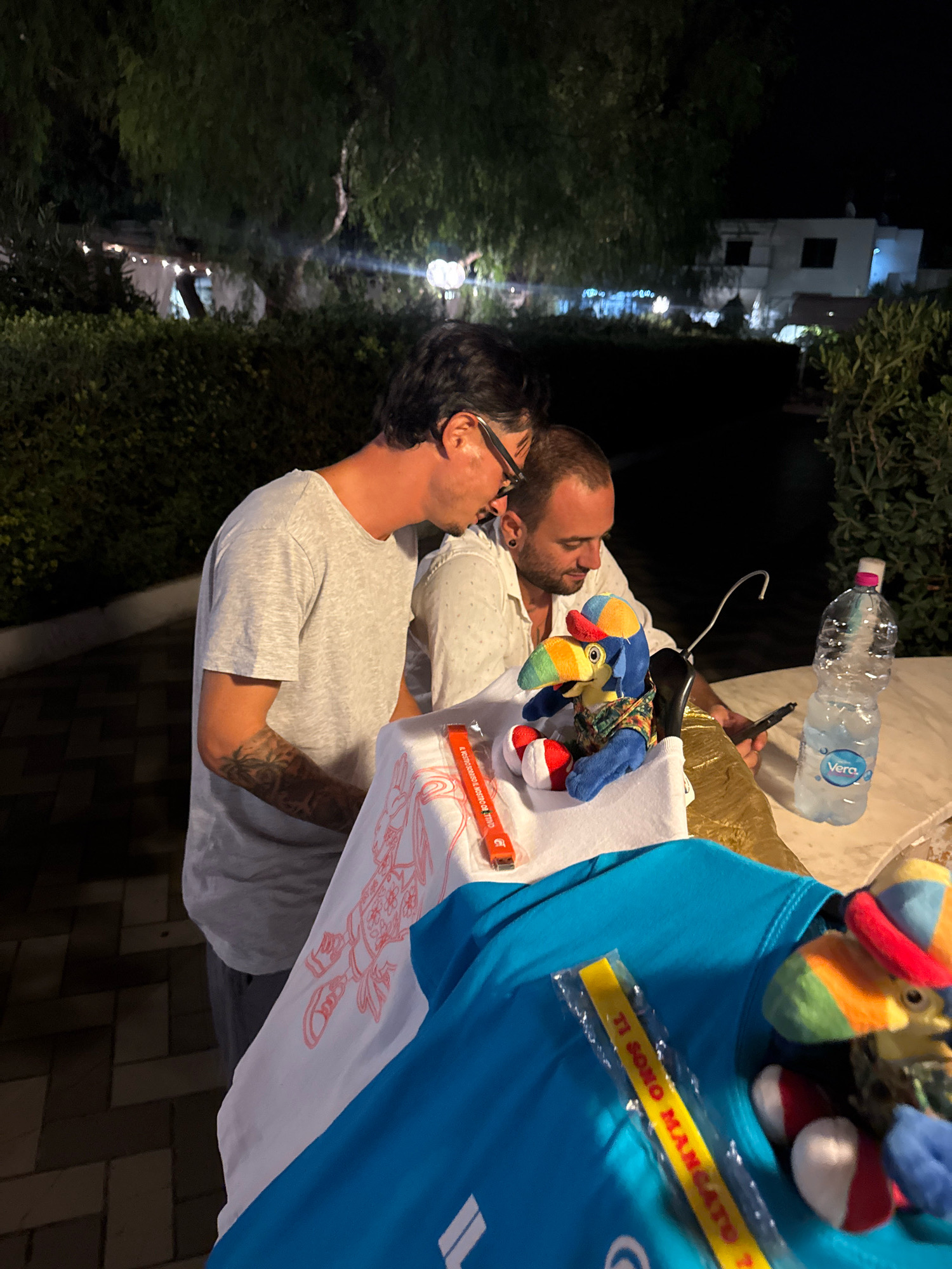 Two men are standing next to a table outdoors at night. The man on the left is wearing a grey t-shirt and glasses. He has tattoos on his left arm. The man on the right is wearing a white shirt with a dotted pattern. He is looking down at a phone or tablet and holding a pen. In front of them on the table are several items, including a plush toy of a parrot wearing a colorful shirt and hat, a white towel with a red graphic print, a bottle of Vera brand water, and a blue t-shirt with a yellow bracelet-like item partially visible. The background shows dark foliage, trees, and some distant lights and buildings, suggesting an outdoor setting in a town or city at night. The man on the right appears to be in the process of signing something with the pen. The location is identified as Isola delle Femmine, Italy.