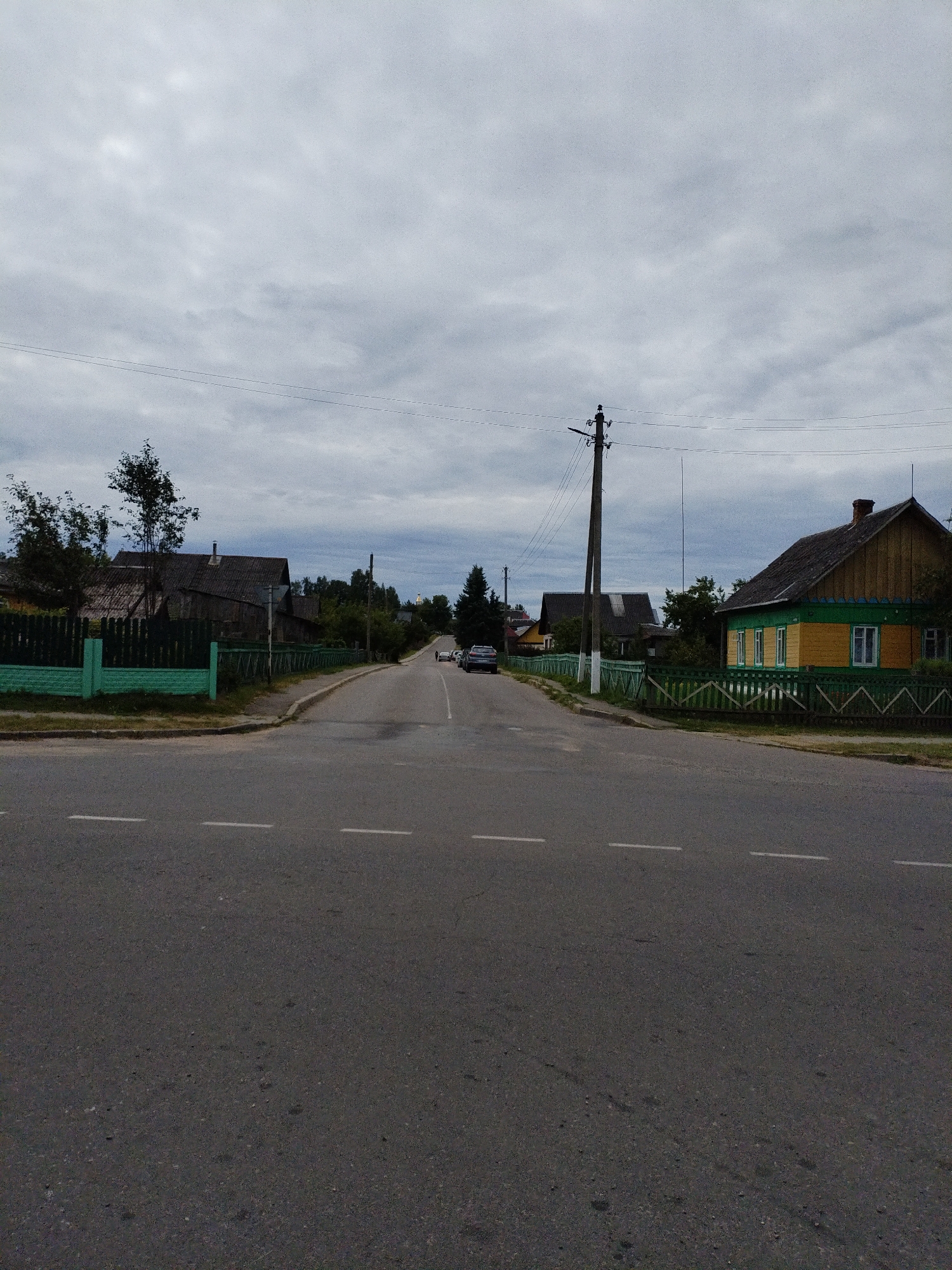The image depicts a rural street scene in Bialynichy, Belarus, under a cloudy sky. The road, asphalt with dashed white lane markings, curves gently to the right and recedes into the distance. On the left side of the road, a green wooden fence runs along the perimeter of several houses with dark roofs. Trees are visible behind the fence. On the right side of the road, a brightly painted yellow and green house with a red-brick chimney is prominent, also enclosed by a green wooden fence. Further down the road on the right, a few cars are parked or in motion, and a power pole with electrical wires stands tall. The environment appears to be a residential area in a village or small town.