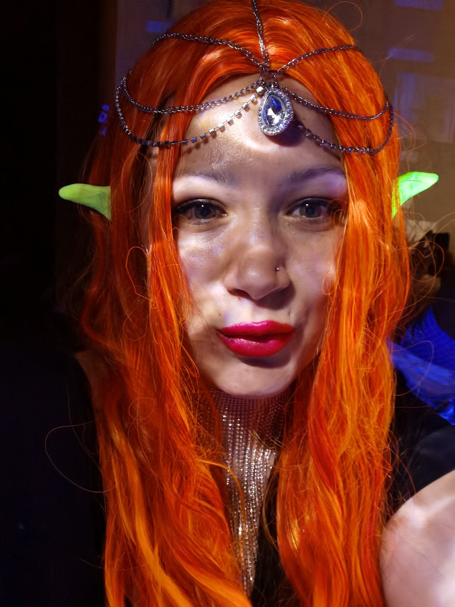 A close-up selfie shows a person with fair skin, dressed as an elf, with bright orange, wavy hair, and pointed, neon green ears. They are wearing a multi-strand silver headpiece with a large, teardrop-shaped jewel on the forehead. The person has dark eyes, a nose piercing, and wears bright red lipstick. They are also wearing a silver, mesh-like necklace. The background is dark, with indistinct shapes and colors, including a patch of blue. The location and city of Chaikovsky, Russia cannot be determined from this image alone.