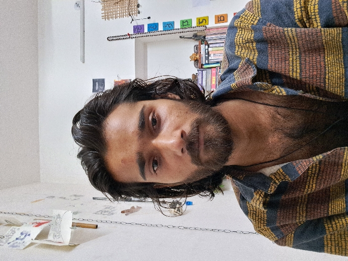 The image shows a young man with dark hair and a beard wearing a blue, yellow, and red striped shirt. He is looking directly at the camera. His chest hair is visible through the open collar of his shirt. The background shows a white wall with a chain hanging down on the left side of the frame. The image was taken in Meerut, India, located at 29.009° N latitude and 77.756° E longitude. The image appears to have been taken in a home or an apartment, as there are bookshelves visible in the background, containing both books and various knick-knacks. The man has a serious expression on his face. He is not interacting with anyone in the image. The lighting appears to be somewhat dim.