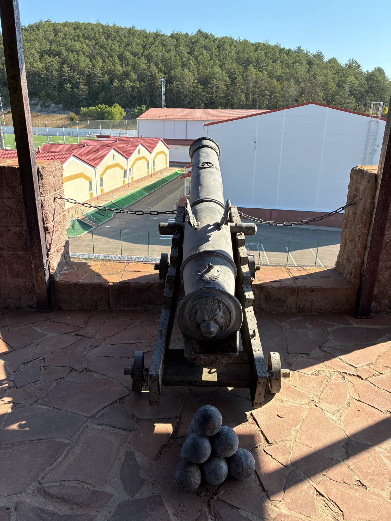 An antique cannon is positioned on a stone patio overlooking a sports complex and a densely forested hillside under a clear blue sky. The cannon, made of dark metal, rests on a carriage with visible wheels and a decorative lion's head embossed on the muzzle. A pile of cannonballs sits in front of it. The patio is paved with irregular flat stones, with sunlight casting strong shadows. Beyond the patio, a chain-link fence separates the area from a sports field with a running track. In the background, several yellow buildings with red roofs are visible, followed by a large white building and the green expanse of the forest. The scene is likely located in Abrau-Dyurso, Russia, a region known for its picturesque landscapes and historical sites. There are no people or animals present in the image, and no activities are taking place.
