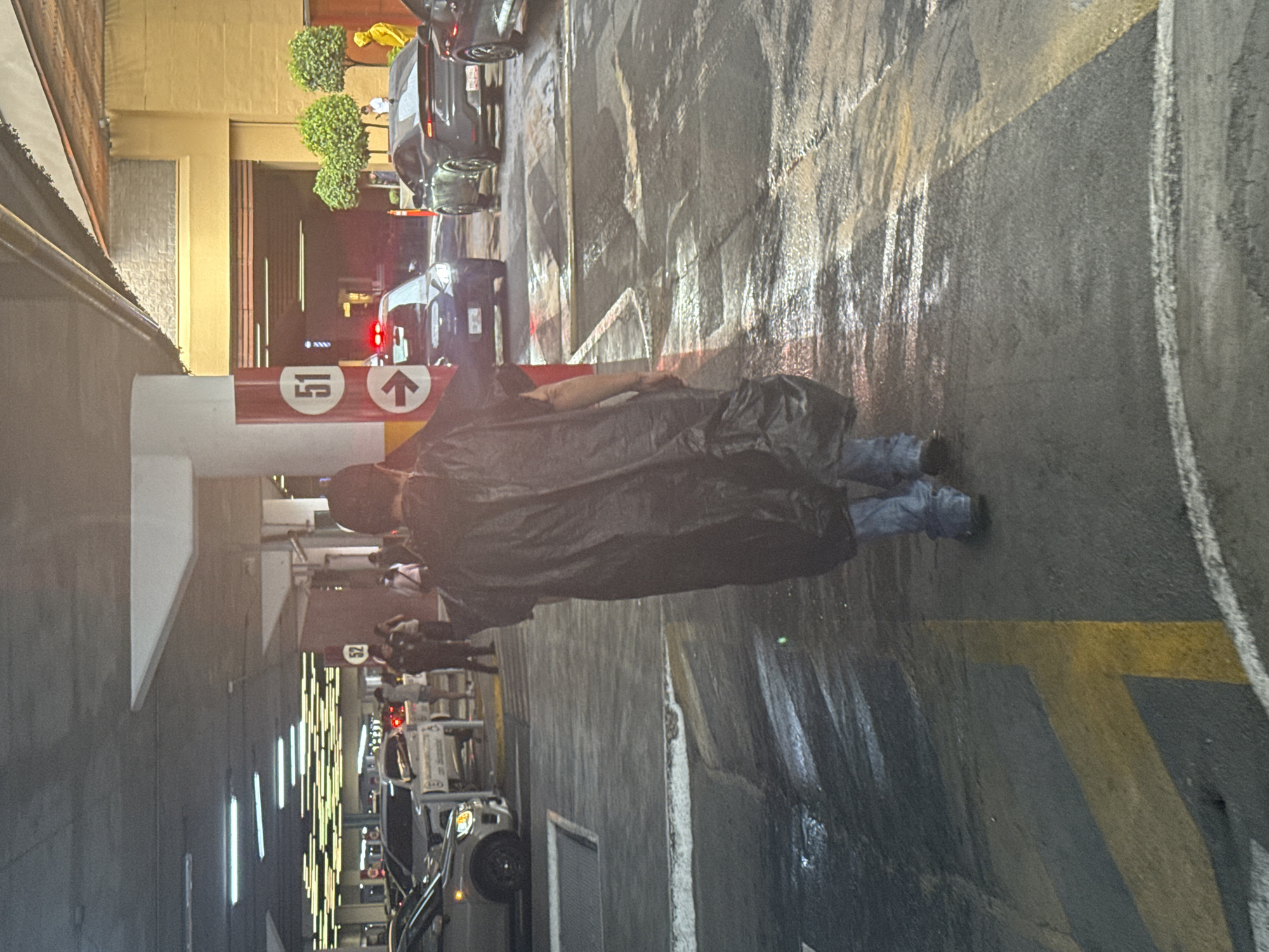 The image captures a rainy scene inside a parking garage in Puebla, Mexico. A person wearing a black raincoat is walking on the wet asphalt floor. Cars are parked nearby, and other people are visible in the background near the entrance/exit of the garage. Signage indicating level "15" is visible on a pillar.