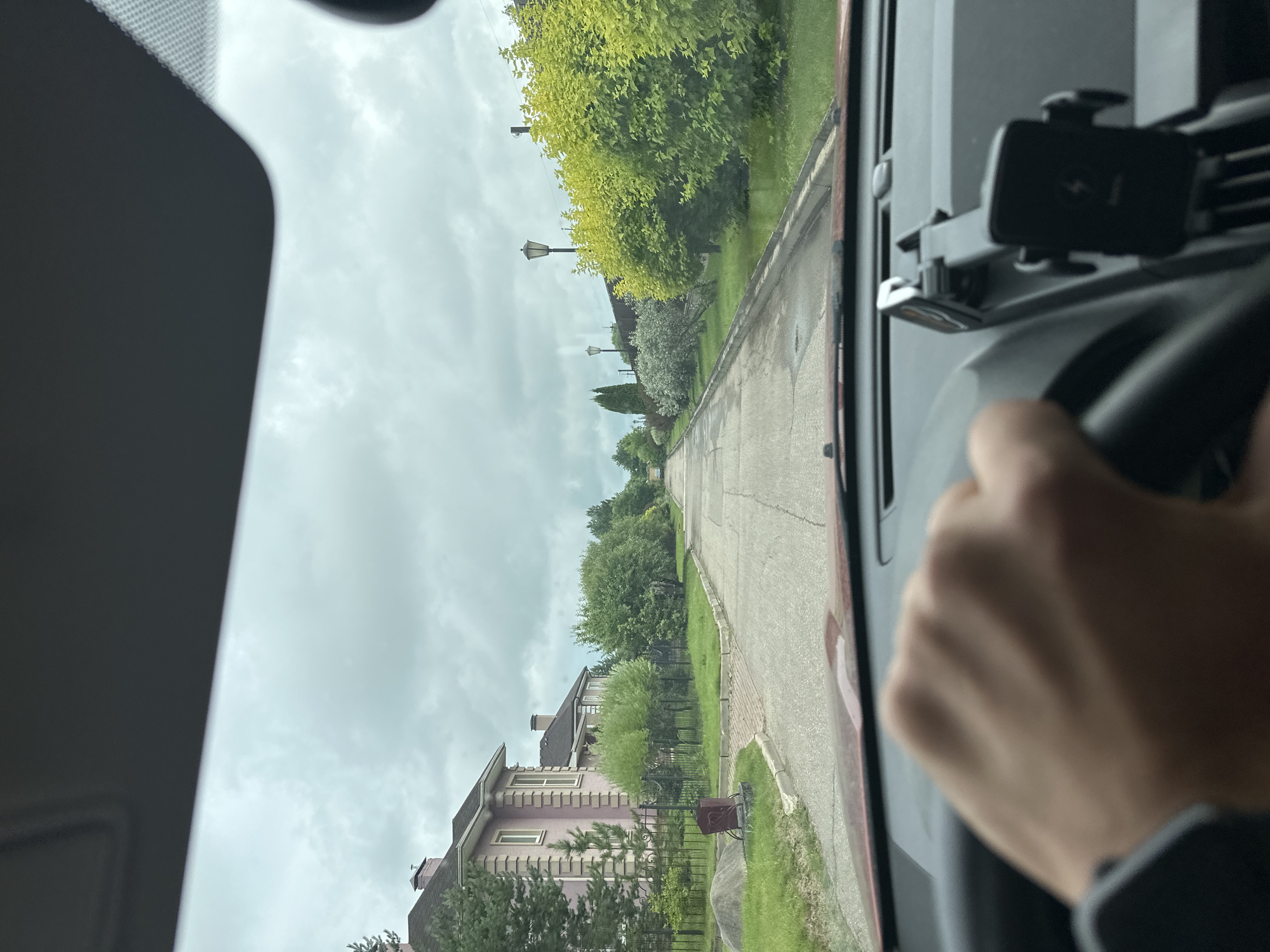 The image shows a view from inside a car driving down a suburban street. The road is paved and appears to be a quiet residential area with houses on either side, characterized by manicured lawns and lush greenery. The sky is overcast, suggesting a cloudy day. In the foreground, a person's hand is visible on the steering wheel. The car is equipped with a phone mount on the dashboard. The houses are of varying architectural styles, some with visible chimneys. There are streetlights along the road.