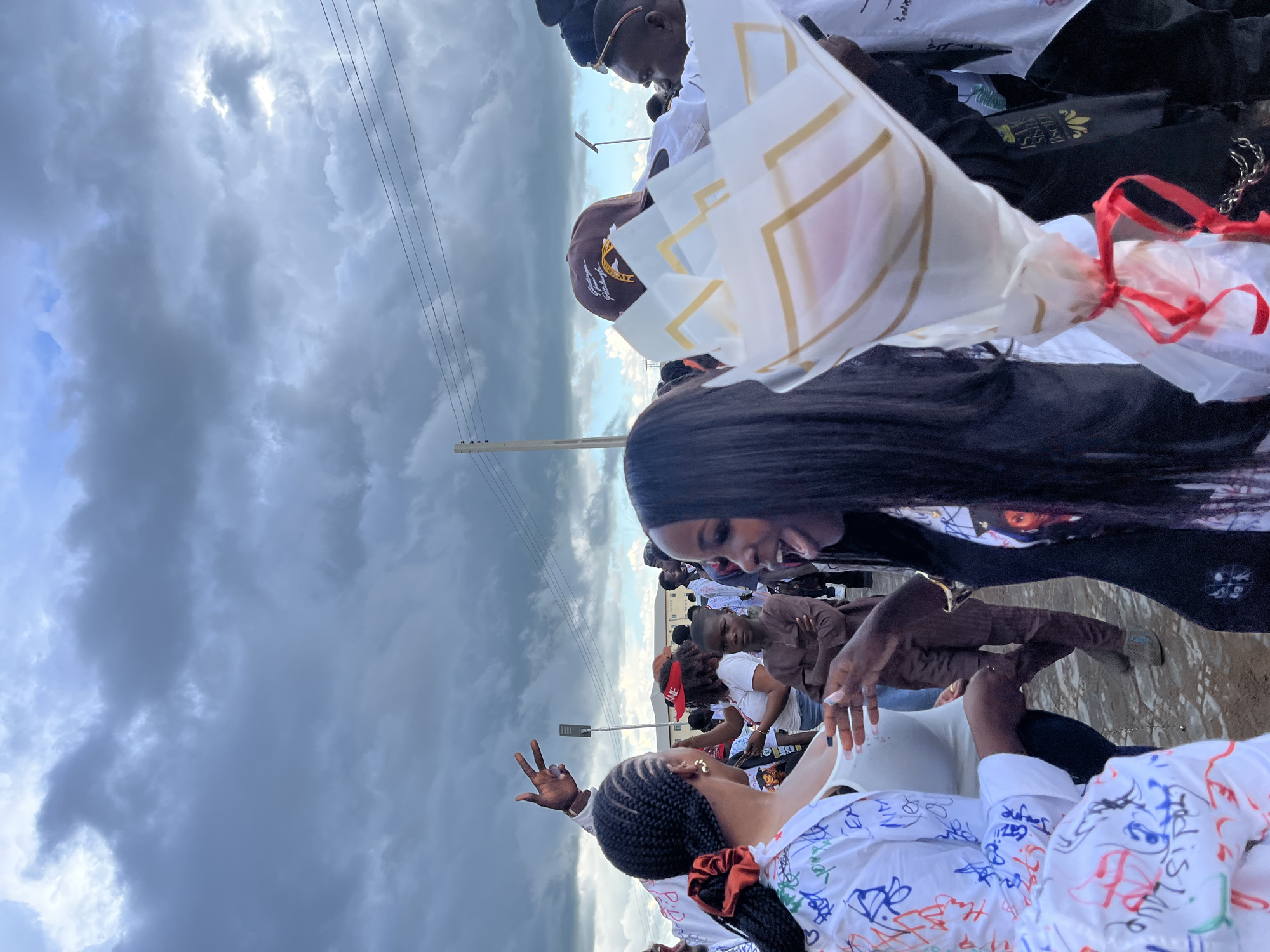 In Rafin Par, Nigeria, an outdoor gathering is underway, characterized by a crowd of individuals under a dramatic, overcast sky. The foreground is dominated by two women engaged in conversation. The woman on the left, with braided hair secured by a red tie, wears a white shirt adorned with colorful scribbles. Her companion on the right, with long, dark hair, faces away from the viewer, her mouth open as if speaking animatedly, and holds a bouquet wrapped in white paper with gold accents, tied with a red ribbon. Behind them, other attendees are visible, some in white attire, and a young boy in traditional brown clothing stands with his arms crossed. The background features a street light, power lines, and buildings, suggesting an urban or semi-urban environment. The overall atmosphere appears festive, despite the dark clouds overhead, hinting at a celebration or social event.