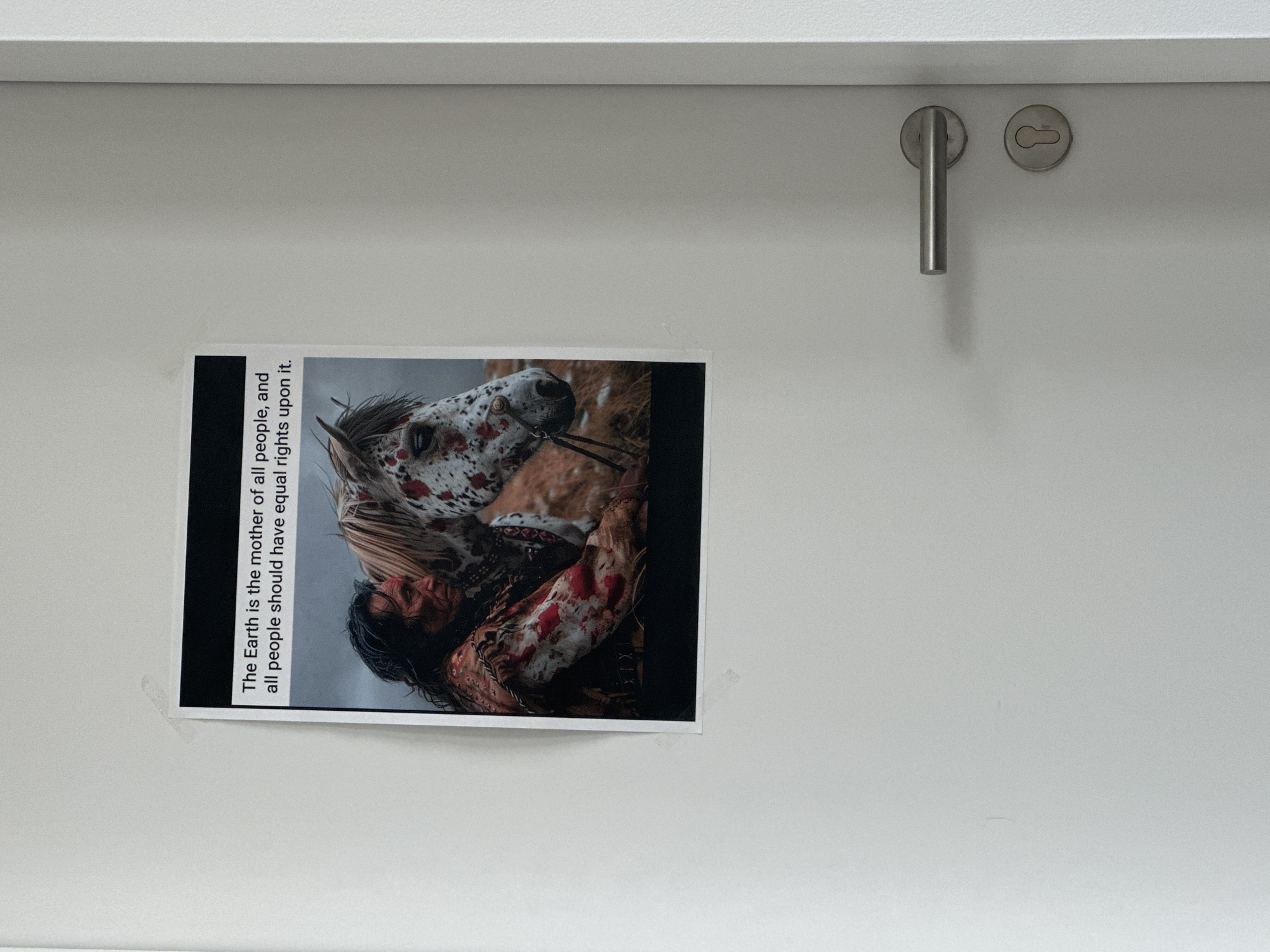 An image depicts a poster on a white door. The poster features an illustration or photograph of a Native American individual embracing a horse. The text on the poster reads, "The Earth is the mother of all people, and all people should have equal rights upon it." The door is equipped with a silver doorknob and a keyhole. The scene is indoors, with the door appearing to be a typical interior door. There is no information to suggest this image is located in Krefeld, Germany.