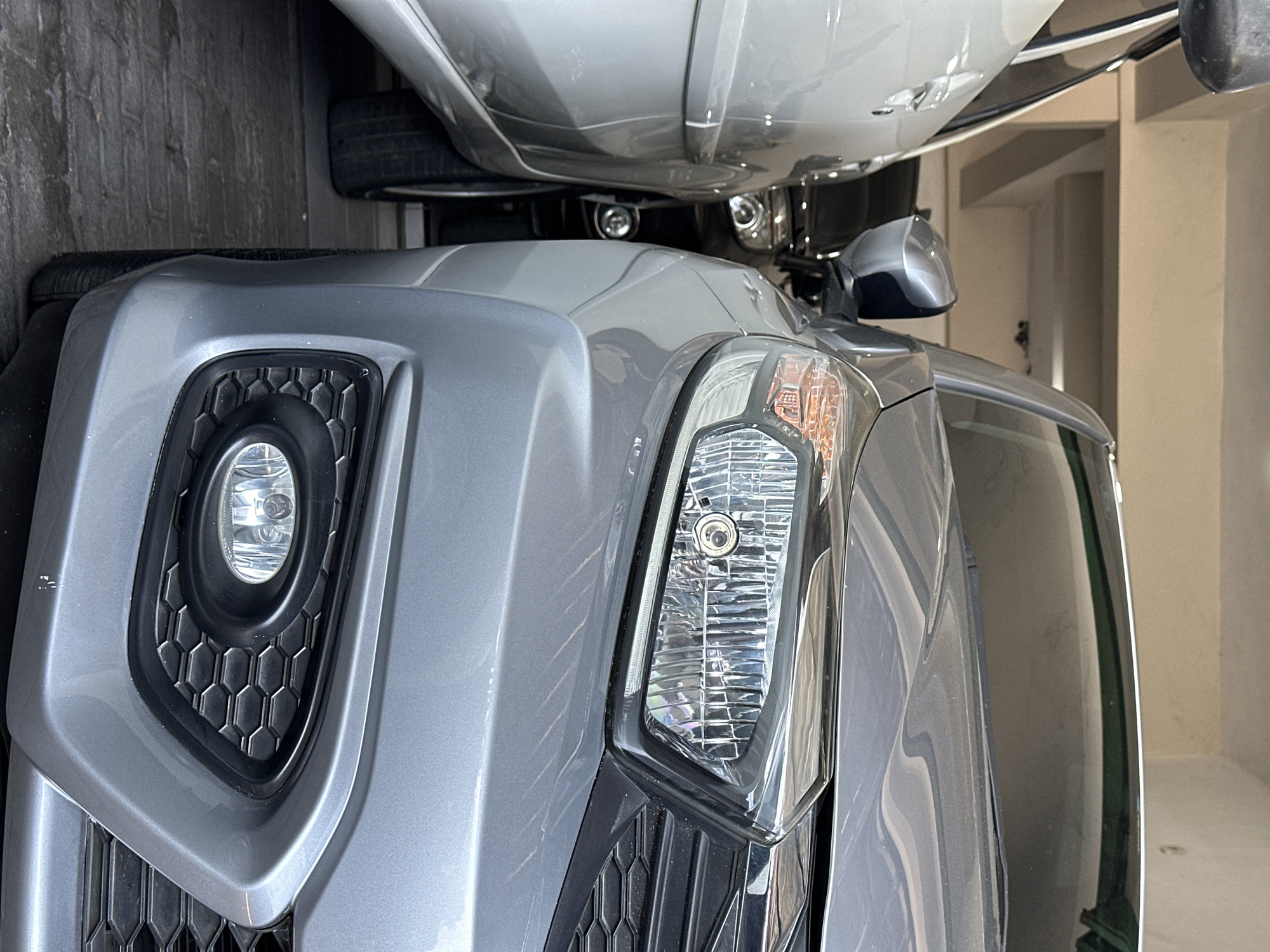 The image shows the front right corner of a silver car parked in what appears to be a residential garage or covered parking area. The car's headlight and fog light are visible, along with a portion of the grille and bumper. To the left of the silver car, another vehicle, possibly also silver or white, is parked. In the background, a dark-colored car is partially visible, parked behind the silver car. The environment is dimly lit, suggesting an indoor setting with concrete flooring and a visible ceiling structure. The specific location of Bulakrejo, Indonesia, cannot be determined from the visual information provided in the image.