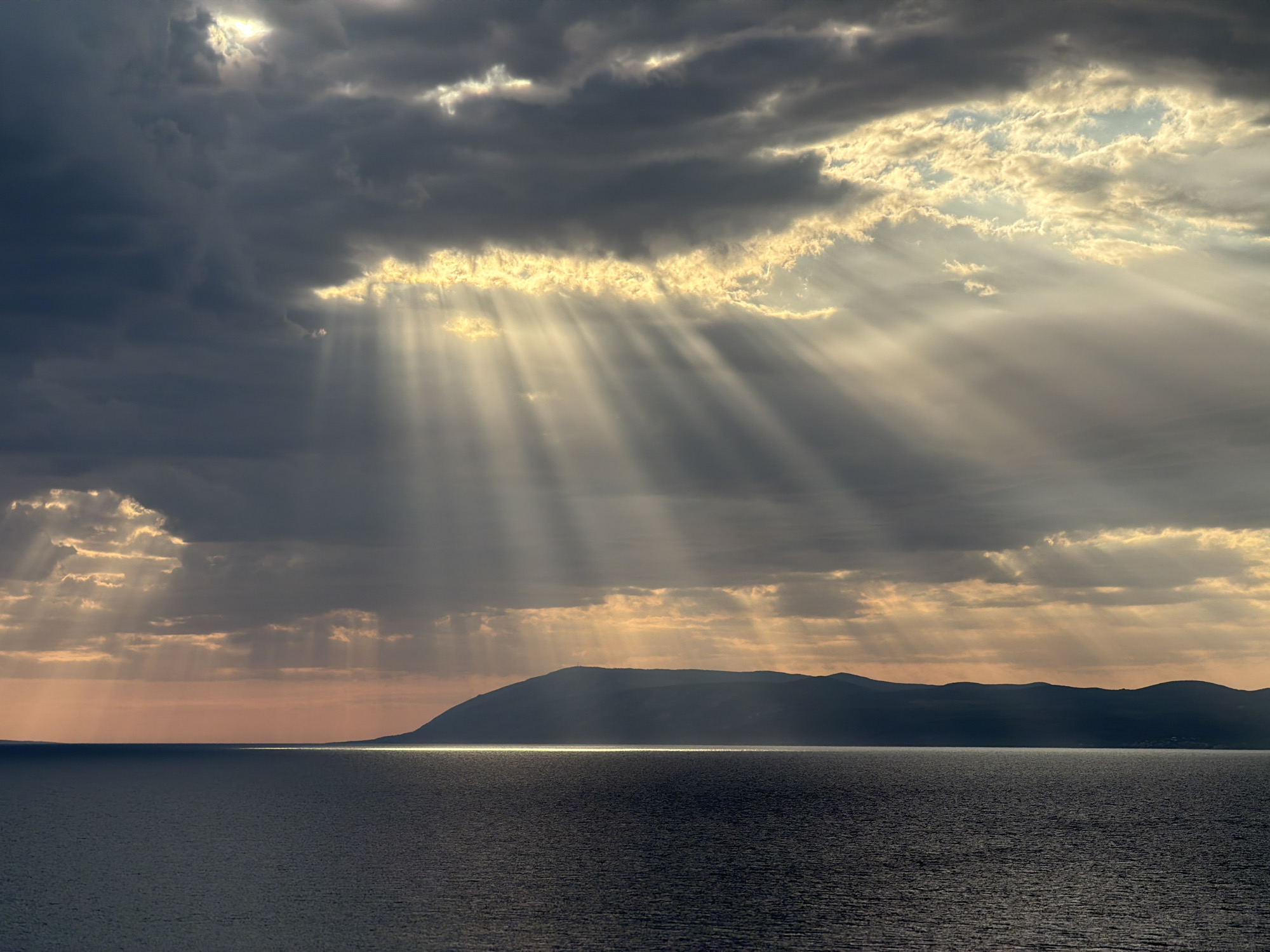 The image depicts a dramatic seascape with heavy, dark clouds partially obscuring the sky. Shafts of sunlight break through the clouds, illuminating the scene with rays of light. The foreground is dominated by a dark, rippling sea. In the distance, a silhouetted mountainous coastline stretches horizontally across the horizon, with a bright reflection of the sun visible on the water just in front of it. The sky transitions from dark gray clouds to lighter, illuminated patches where the sun is breaking through, casting warm hues of orange and yellow on the horizon. The overall impression is one of a moody, atmospheric landscape, possibly taken at sunrise or sunset. The location is identified as Općina Podgora, Croatia.