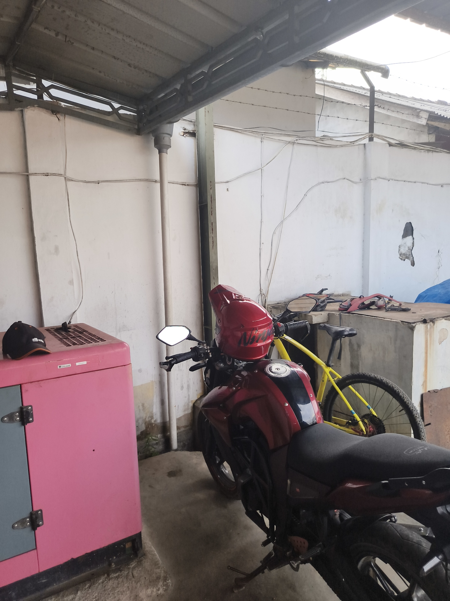 A red motorcycle with a red helmet is parked in an outdoor area next to a pink generator. A yellow bicycle is parked behind the motorcycle. The scene appears to be a storage or maintenance area, with white walls and a corrugated metal roof overhead. The ground is concrete, and there is some debris present. The image is likely taken in the city of Pangkalpinang, Indonesia, given the presence of a generator, which are commonly used in areas with unreliable power.