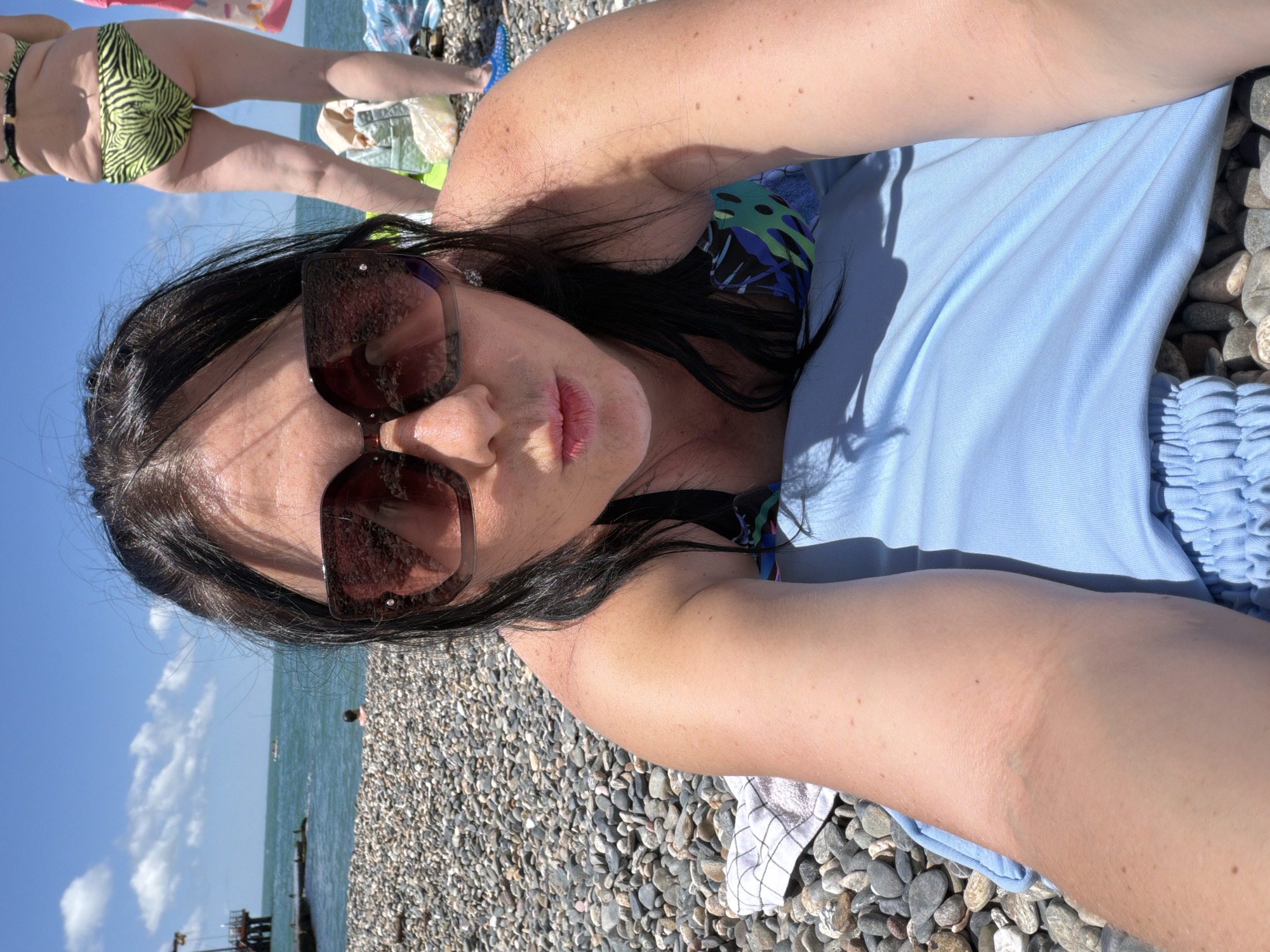 A woman in a bikini lies on a pebble beach on a sunny day, likely in Sochi, Russia, given the presence of a pier and the Black Sea. She is wearing large, dark sunglasses and has dark hair. A second woman in a lime green and black striped bikini is standing in the background, facing away from the camera. The beach is covered in smooth, gray pebbles. The sea is visible behind the woman, with a small boat in the distance. The sky is bright blue with scattered white clouds. The woman in the foreground is lying on a light blue towel or blanket.