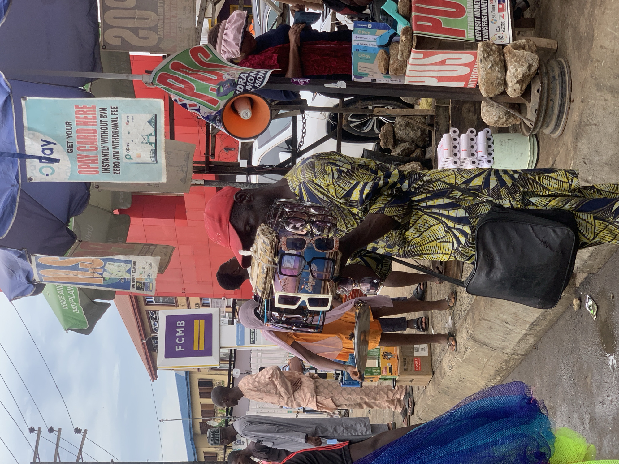 The image depicts a street scene in Ikorodu, Nigeria. A sunglasses vendor wearing a yellow patterned shirt and red cap stands on the sidewalk, displaying sunglasses on a tray. Several people are standing or walking nearby. Advertisements and signs are prominently featured, including those for "Opay Card Here," "FCMB," and "Western Money Transfer." The scene is set against a backdrop of buildings. The environment appears to be a busy commercial area, with various goods and objects scattered around. A loudspeaker is also visible.