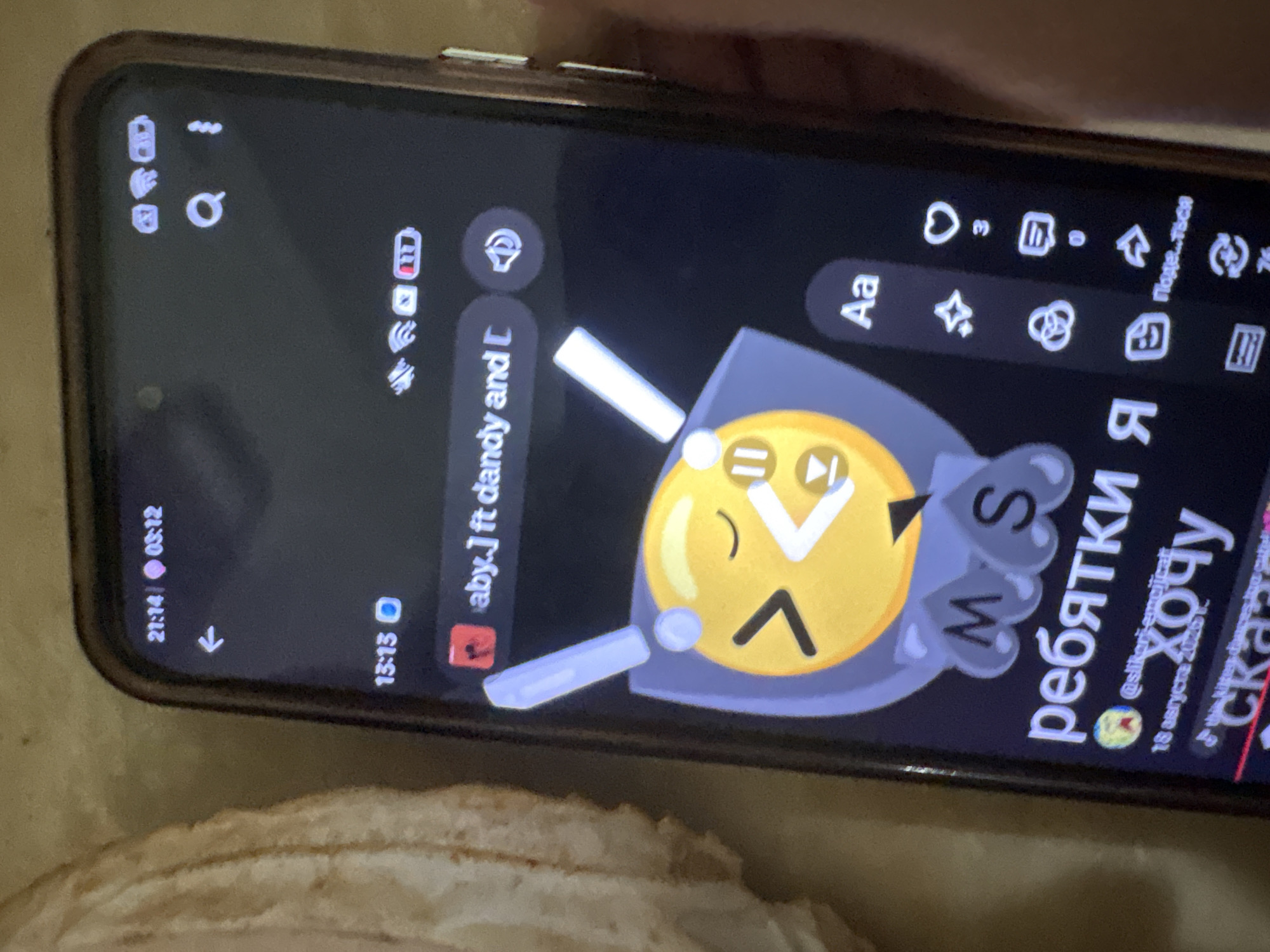 The image is a close-up, slightly blurred photograph of a smartphone screen displaying a social media or messaging application. The phone is held in a person's hand, with a portion of their fingers visible on the right side of the frame. The background appears to be a textured surface, possibly fabric, in a neutral tone.
On the phone screen, there is a status bar at the top showing the time as "21:14" and "03:12", along with battery and network indicators. Below this, there is a back arrow and a search icon. The main content of the screen features a large, stylized yellow emoji with eyes closed in a smile and what appears to be a musical note or a pause symbol. Surrounding the emoji are stylized gray shapes and letters, including "M" and "S". Below the emoji, text in Russian is visible, reading "ребятки" and "хочу". There is also a username "@slikori-emojicat" and a date "18 августа 2023 г.". Further down, the word "сказ" is partially visible. On the right side of the screen, there are icons for liking (a heart with "3" below it), commenting (a speech bubble with "0" below it), sharing (an arrow), and other interaction buttons.
The photograph was taken indoors or in a controlled environment, as indicated by the artificial lighting and the focus on the phone screen. There is no discernible connection to Ashvem, India, in the image itself.