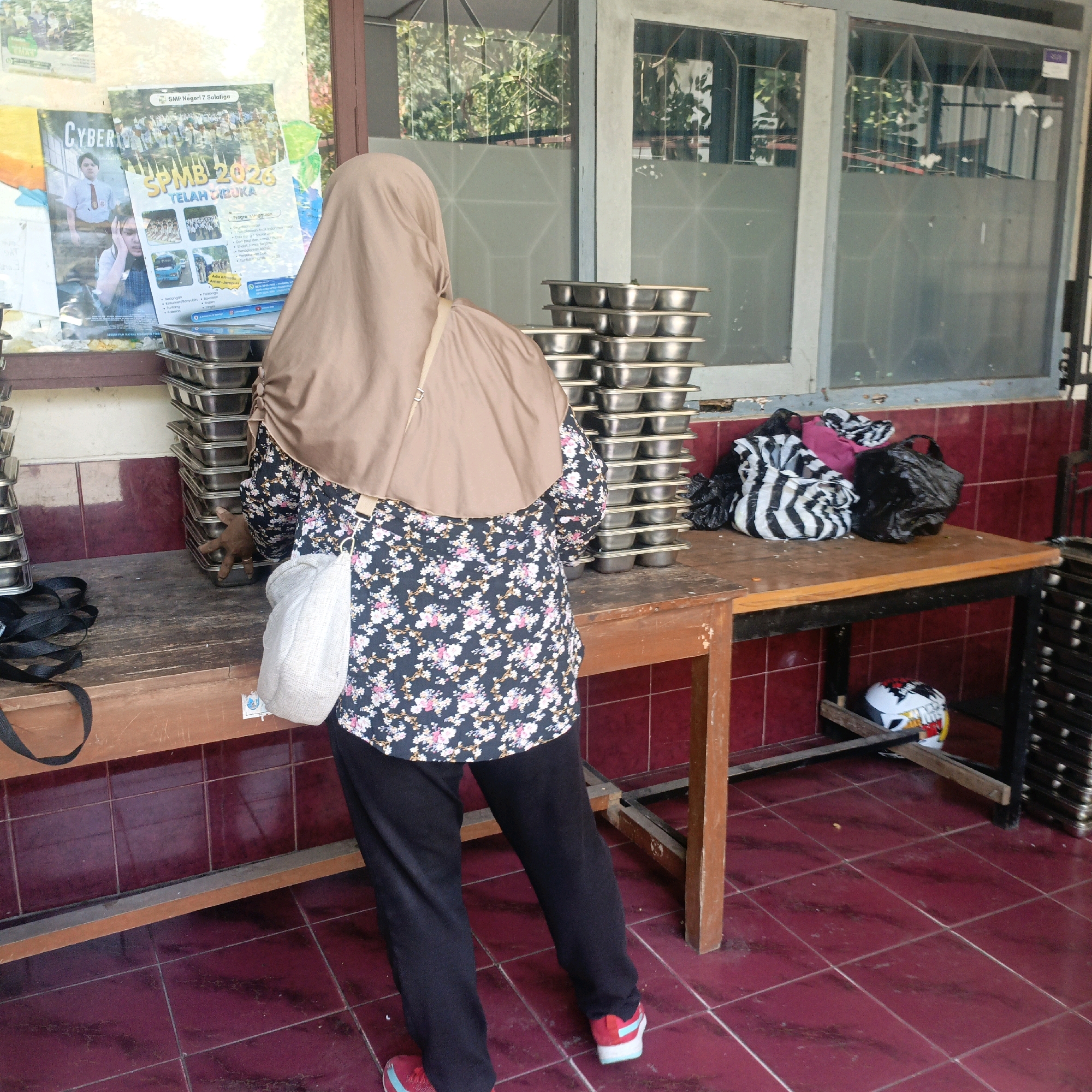 A person wearing a brown hijab and a floral print shirt is standing in front of stacks of metal trays. The person is holding a white bag. The trays are on a wooden table. There are also bags of trash on the table. In the background, there is a poster on the wall and windows with reflections of trees. The floor is tiled in a reddish-brown color. The scene appears to be an outdoor or semi-outdoor area, possibly a school or community center, given the poster which advertises "SPMB 2026" for "SMP Negeri 7 Salatiga," indicating it is located in Salatiga, Indonesia.