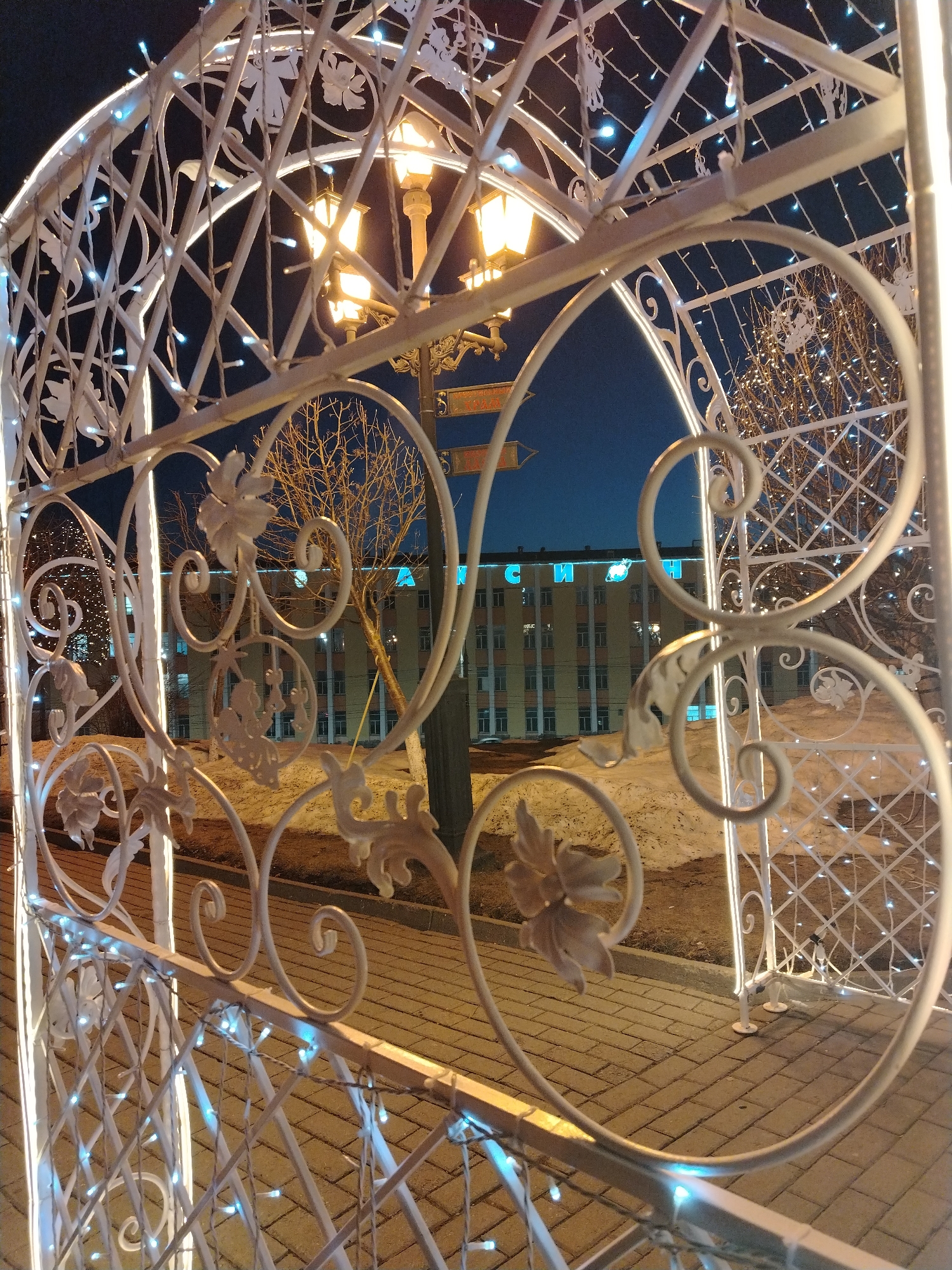 The image shows a decorative white metal archway decorated with illuminated string lights. The archway is located on a brick-paved walkway. Through the archway, a building is visible with a sign in Russian, "Я ИЖЕВСКАЯ," which translates to "I am Izhevsk." A bare tree stands in front of the building. A street lamp is visible behind the archway. The scene appears to be at dusk or night. The location is Izhevsk, Russia.