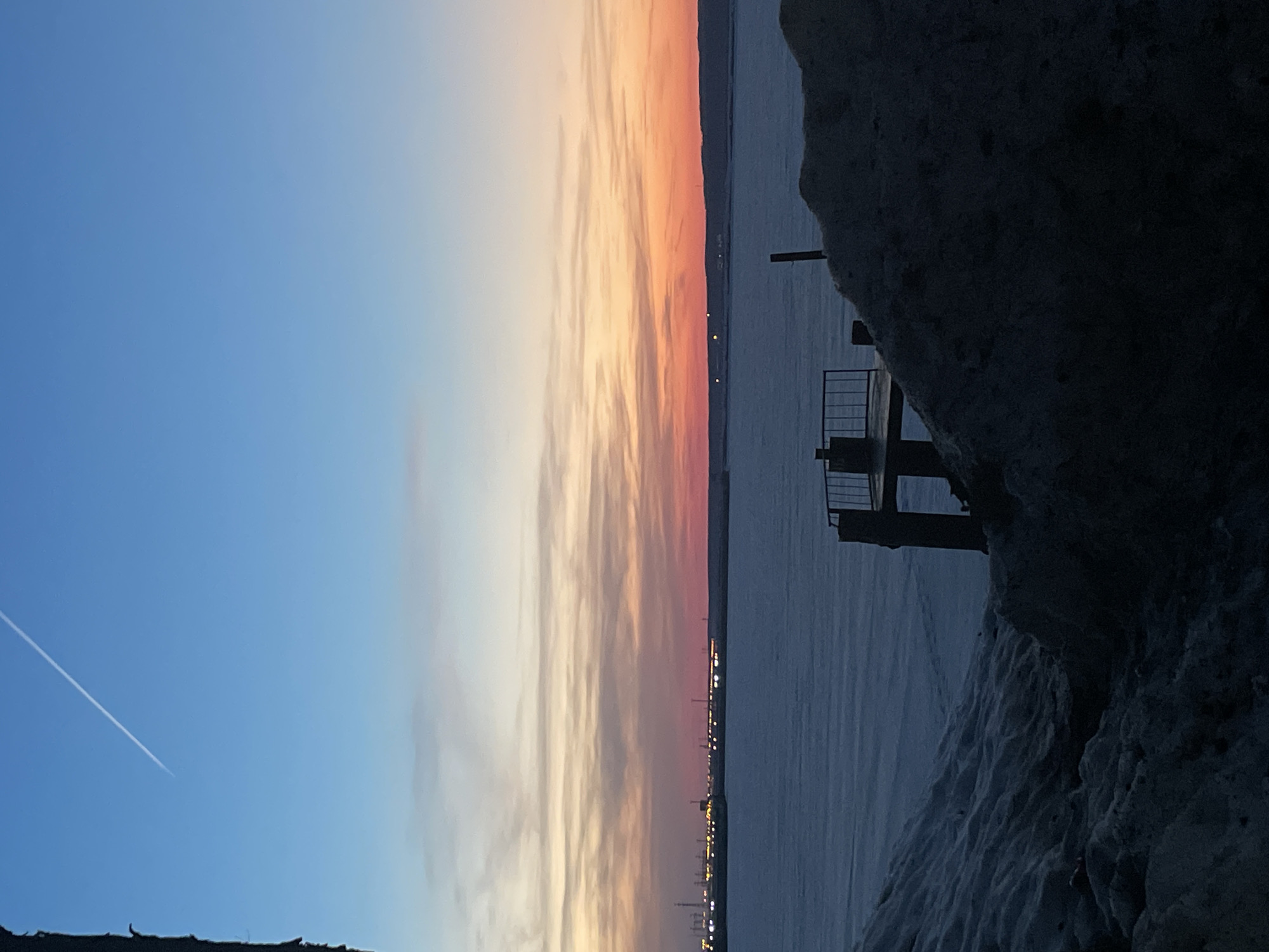 The image depicts a winter scene at either dawn or dusk, overlooking a frozen body of water near Chaikovsky, Russia. The sky transitions from a light blue at the zenith, featuring a prominent white aircraft contrail diagonally traversing from the upper left, to a horizontal band of orange, pink, and light blue layered clouds along the horizon. A broad expanse of ice or snow covers the water, extending across the midground. On the distant left horizon, a cluster of industrial lights and indistinct structural silhouettes are visible, while scattered smaller lights extend further right along the horizon. The foreground is dominated by irregular mounds of snow and ice; a lighter, textured mound occupies the lower left, and a darker, less defined mound occupies the lower right. Between these mounds, a dark, weathered pier or small platform with a metal railing stands on two vertical piles. No individuals or specific activities are visible.