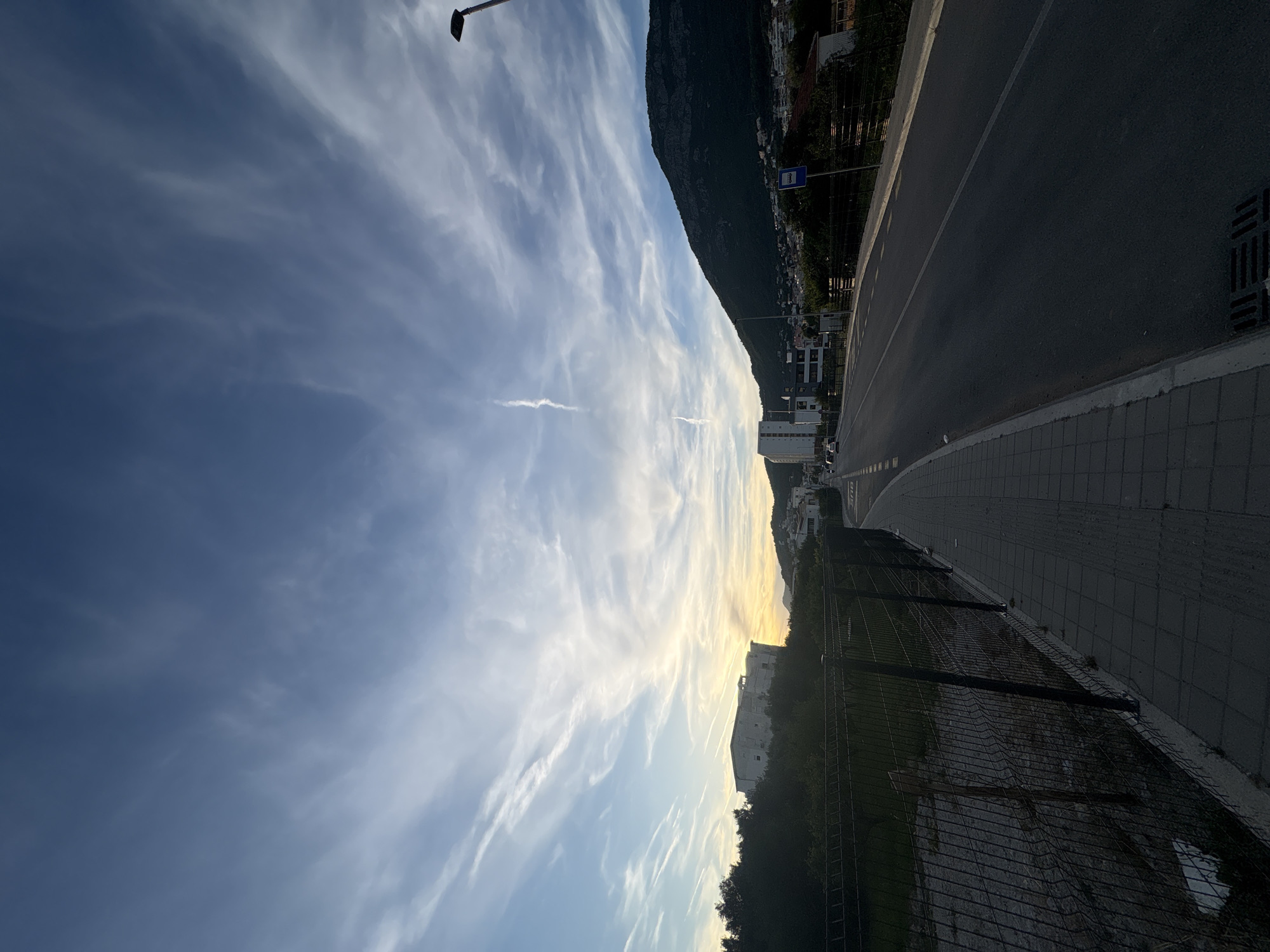 This image captures a scenic view of a road leading towards a mountain in Bar, Montenegro. The sky is a dramatic canvas of blue with wispy white clouds stretching across it, hinting at a sunset or a transition in weather. The road itself is asphalt, marked with white lines, and a paved sidewalk runs alongside it, separated by a dark metal fence.
On the left side of the road, beyond the fence, is a patch of dry, rough terrain with some sparse vegetation. In the middle ground, several modern buildings, primarily residential or commercial, are visible, with a distinctive white building standing out among them. The architecture suggests a contemporary urban or suburban setting.
To the right, the imposing dark silhouette of a mountain dominates the landscape, its peak partially obscured by the atmospheric conditions. The lower slopes of the mountain are dotted with more buildings and lush greenery. A street lamp is visible on the right side of the image, indicating the presence of street lighting. A bus stop sign with a blue icon is also present.
The overall impression is one of a tranquil evening in a coastal Montenegrin city, where nature and human habitation coexist. The lighting suggests it's either late afternoon or early evening, with the sun casting a warm glow on the horizon, visible behind the buildings and trees.