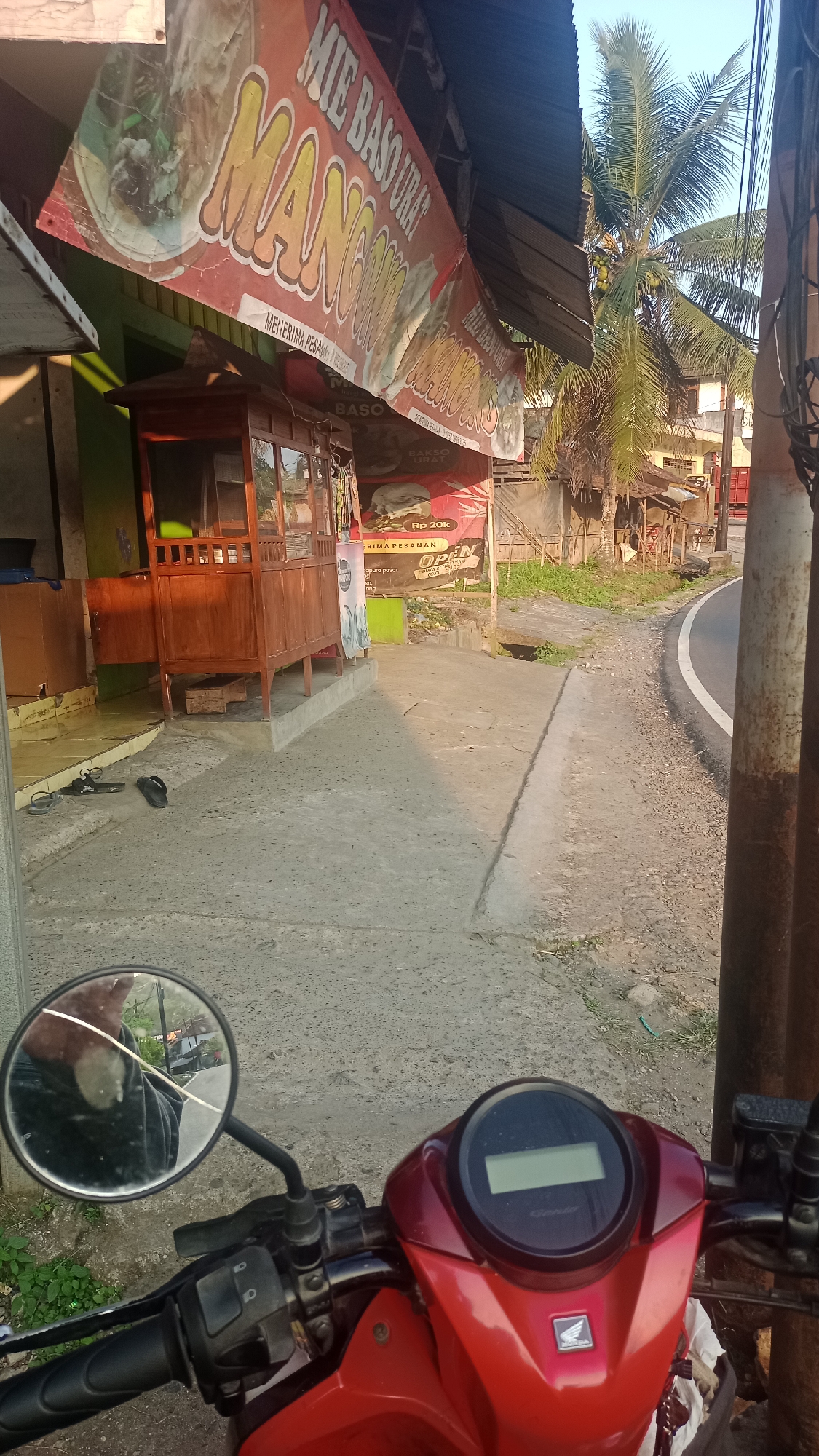 The image captures a street scene in Cogreg, Indonesia, with a focus on a roadside food stall. A red motorcycle is parked in the foreground, with its handlebars and instrument panel visible, indicating the perspective of someone seated on the bike. A cracked rearview mirror is attached to the handlebars, reflecting a person's arm and shoulder.
To the left of the motorcycle, a wooden food stall with glass windows and a covered roof stands on a concrete platform. Above the stall, a red banner with white and yellow lettering advertises "MIE BASO URAT MANG NONO," suggesting it sells beef tendon noodles and meatballs. Below the banner, smaller signs display prices, including "Rp 20k." Two pairs of flip-flops are on the ground near the stall.
The scene is illuminated by the warm light of what appears to be late afternoon or early morning sun, casting long shadows across the concrete pathway. A paved road curves to the right of the pathway, bordered by gravel and sparse vegetation. In the background, a tall palm tree with coconuts grows beside a few simple buildings, some with corrugated metal roofs, typical of residential or small commercial structures in the area. The overall impression is of a casual, everyday street view in a local Indonesian town.