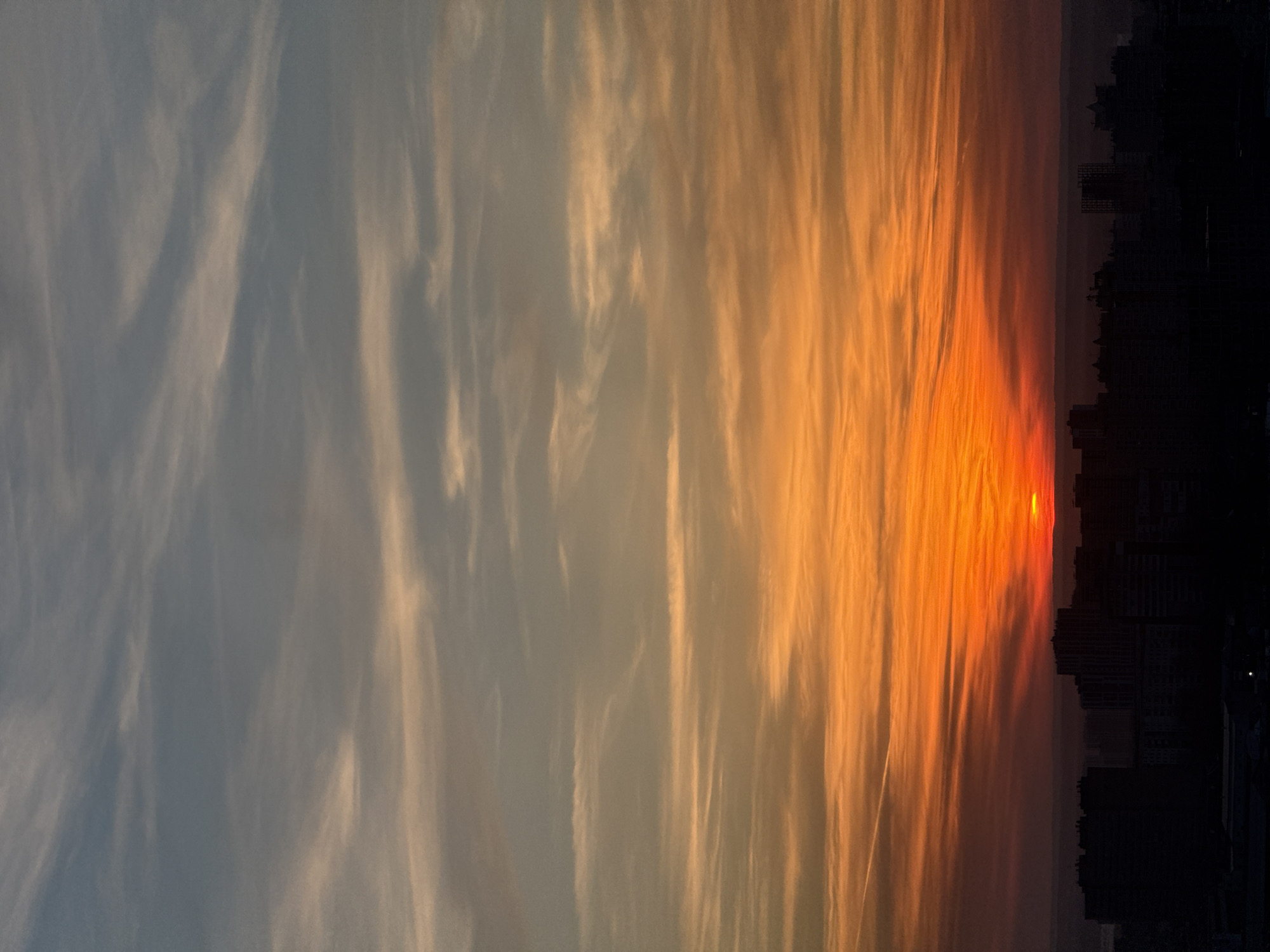This image depicts an urban sunset or sunrise over the city of Perm, Russia, observed from an elevated vantage point. The sky occupies the majority of the frame, displaying a pronounced horizontal gradient. The lowest portion of the sky, near the horizon, is dominated by intense hues of dark orange and red, where the sun's lower limb is visible as a bright, glowing disc, partially obscured by the distant urban silhouette. Above this, the sky transitions through vibrant oranges and yellows, gradually fading to a lighter blue-grey at the top of the frame. Numerous wispy, elongated clouds are present across the entire sky, illuminated by the sun, creating striated patterns. A single, distinct white airplane contrail extends diagonally in the upper-left region of the orange sky. The lower quarter of the image is occupied by the dark silhouette of a dense city skyline. Multiple multi-story buildings of varying heights, consistent with residential or commercial structures, are visible as featureless, black forms against the bright horizon. No individuals, specific actions, or interactions are discernible within the frame. The scene captures the atmospheric conditions typical of dawn or dusk.