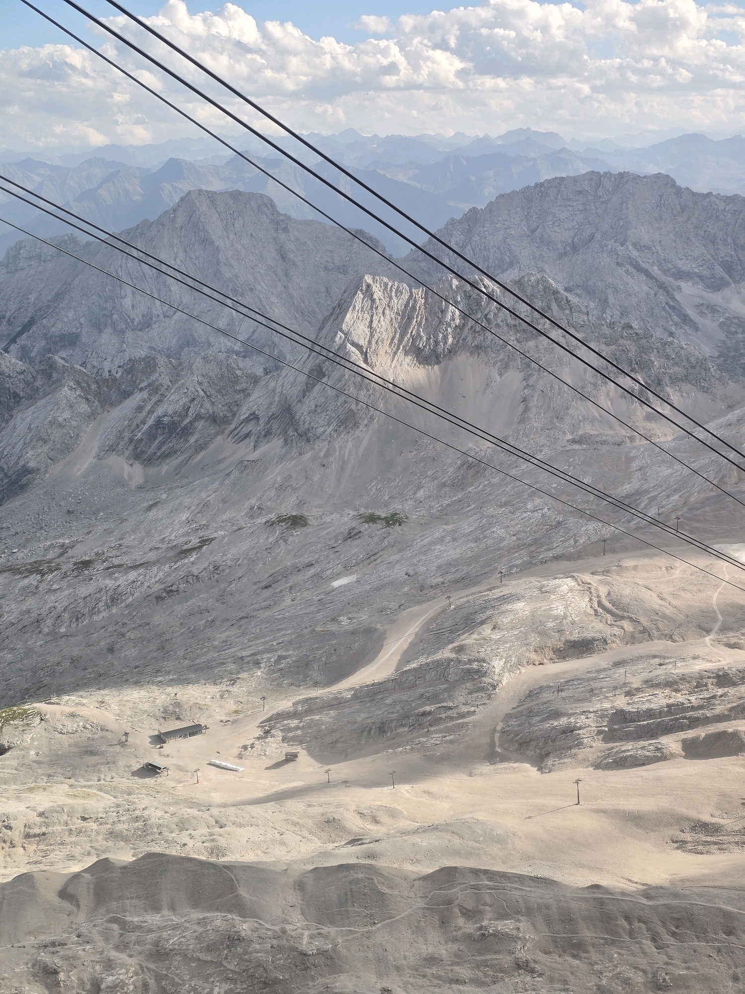 This is an aerial view of a mountainous landscape near Grainau, Germany, likely taken from a cable car. The foreground is dominated by a barren, rocky terrain with sparse vegetation and several structures that appear to be part of a ski resort or mountain station, including buildings and ski lifts. In the mid-ground, the landscape transitions into a rugged mountain slope with visible geological formations and ski trails. The background features a series of layered mountain ranges extending into the distance, under a sky filled with scattered clouds. Multiple thick cables, presumably part of the cable car system, stretch diagonally across the frame from the top left to the bottom right, partially obscuring the view of the mountains. The overall impression is of a high-altitude, rugged natural environment with human infrastructure adapted to the mountainous terrain.