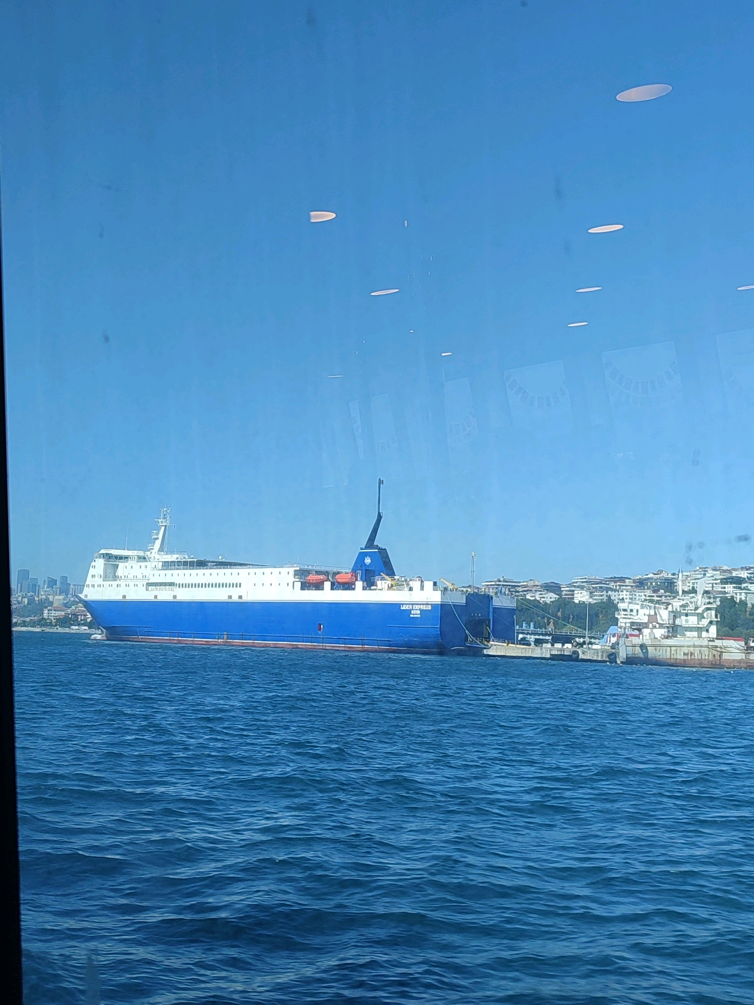 A large blue and white ferry, identified by text on its side as "MIGIR EXPRESS," is docked in the Bosphorus Strait, with the Üsküdar district of Istanbul, Turkey, visible in the background. The ferry is the dominant subject, positioned centrally and occupying a significant portion of the frame. Its hull is predominantly blue, with a white superstructure. Several lifeboats are visible along the side of the ferry.
The Üsküdar skyline is a secondary element, showing a dense collection of buildings with a mix of architectural styles, including modern apartments and older structures. Some greenery is interspersed among the buildings, suggesting hills or parks. The water in the foreground is a deep blue, with moderate ripples indicating a gentle current. The sky above is clear and a bright blue, with a few faint, elongated light reflections on the surface of what appears to be a window or protective covering.
The overall scene is a daytime view of a working harbor or port area. There are no people clearly visible on the ferry or in the immediate vicinity. The image has a slightly hazy quality, possibly due to distance or atmospheric conditions.
