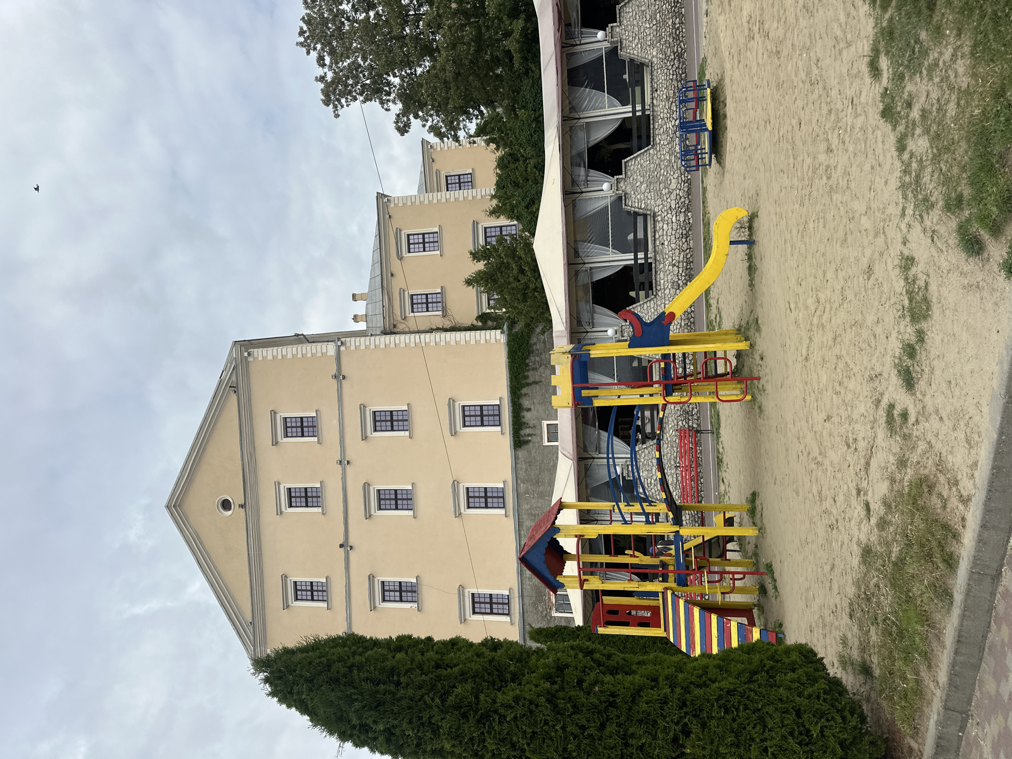 The image displays a playground situated in the foreground, with the eastern facade of Ternopil Castle and an associated restaurant terrace in the midground, all under an overcast sky in Ternopil, Ukraine. The playground occupies a sandy area featuring scattered patches of green grass, bordered by a concrete curb and red brick paving on the left. Play equipment includes a multi-colored (red, yellow, blue) ramp leading to a red and yellow structure with a blue roof on the left, a yellow slide attached to a red and yellow climbing frame in the center-right, and a small blue and yellow merry-go-round on the far right. A tall, slender, dark green coniferous tree stands prominently on the far left.
In the midground, below the castle, a restaurant terrace is visible, characterized by a stone-faced retaining wall with dark railings and a continuous white canvas awning featuring multiple bays with translucent white curtains. The background is dominated by the multi-story Ternopil Castle, presenting a pale yellow stucco facade. It features a distinctive triangular pediment at the apex, containing a small circular oculus window. The facade is punctuated by numerous regularly spaced rectangular windows with dark frames and white sills, some showing dark grilles. White decorative trim highlights architectural elements. A dark, pitched roof and chimneys are partially visible on the right side of the castle. A large deciduous tree with green foliage is partially visible on the far right. The sky is uniformly grey and cloudy. A single small, dark bird is captured in flight in the upper central portion of the sky. No people are clearly visible within the scene.