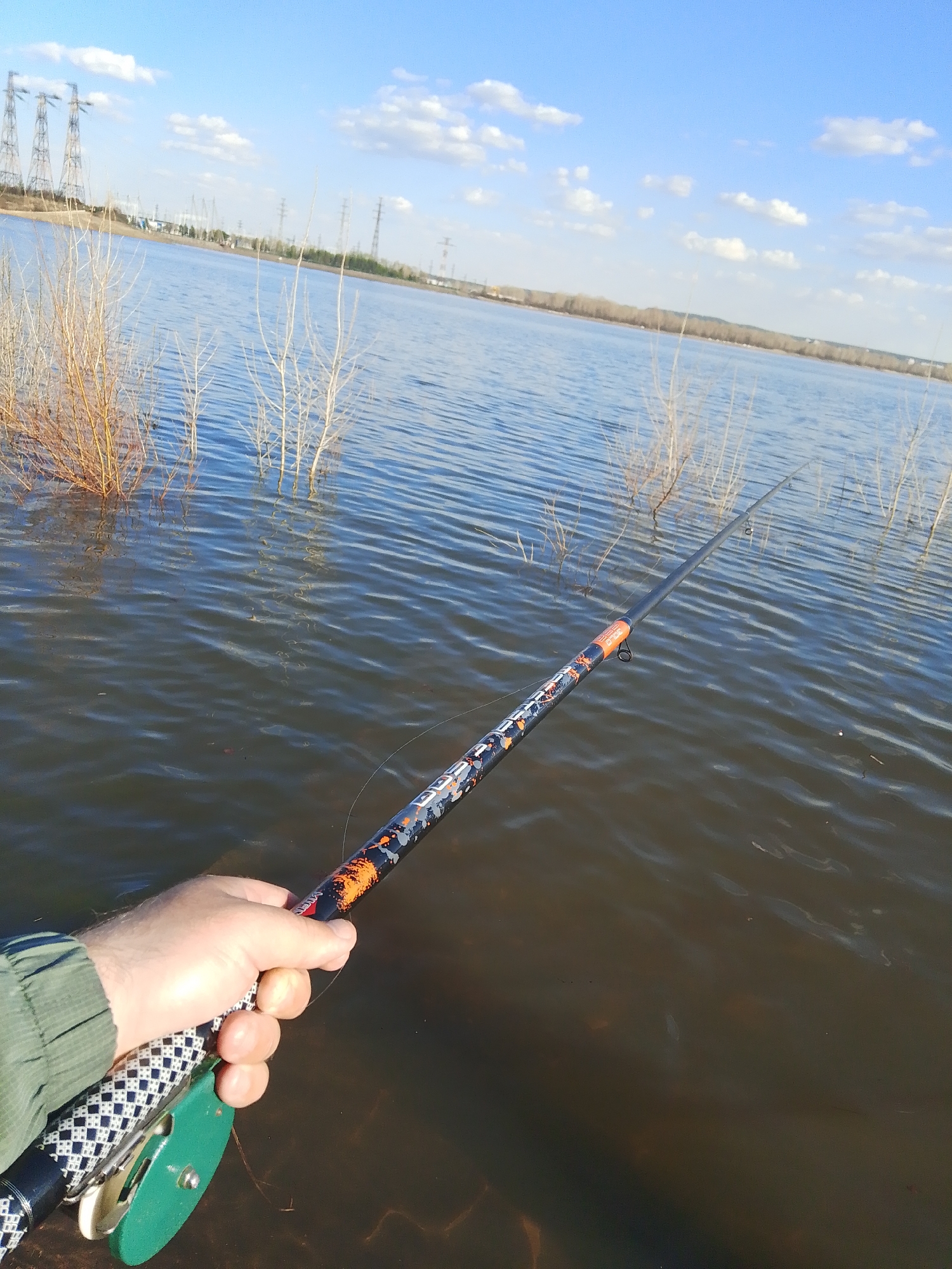 A person is holding a fishing rod over a body of water. The rod is black and orange with a green and white reel. The person's hand is visible, holding the rod. The water is calm with some ripples. In the background, there are bare trees and power lines. The sky is blue with some white clouds. The location is likely the reservoir near Chaikovsky, Russia.