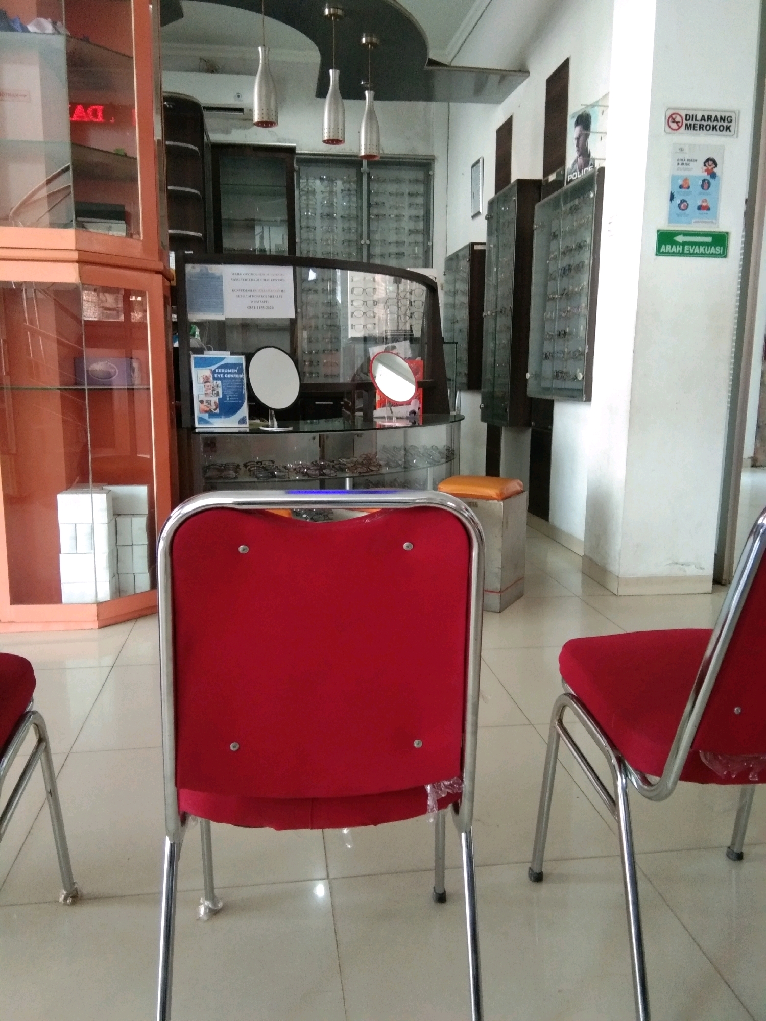 The image depicts the interior of an optical shop, likely located in Kebumen, Indonesia, given the presence of a sign in Indonesian. In the foreground, two red chairs with chrome legs are visible from the back. To the left, a tall, orange-toned display cabinet with glass shelves contains various items, including stacked white boxes. Behind the chairs, a glass display case filled with eyeglasses is positioned on a shelf. Further back, several tall, dark glass display cabinets showcase more eyeglasses. Above these cabinets, a poster is affixed to a white wall. A sign prohibiting smoking ("DILARANG MEROKOK") is also visible on the wall, along with a green directional arrow sign indicating "ARAH EVAKUASI" (evacuation route). The floor is tiled. The overall lighting suggests an indoor environment.