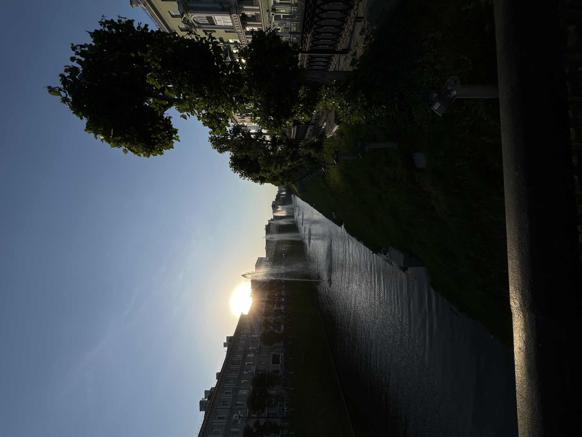 The image captures a water canal scene in Kazan, Russia, with the setting sun directly in the center of the canal. A row of apartment buildings line the lower side of the canal, while trees and an ornate fence line the upper side. Fountains are seen in the canal, silhouetted against the sunlight. A lamppost is situated along the upper edge of the canal and a large, dark, vertical post is in the lower right corner of the frame. The sky is a gradient of light blue. The overall scene conveys a serene urban environment.