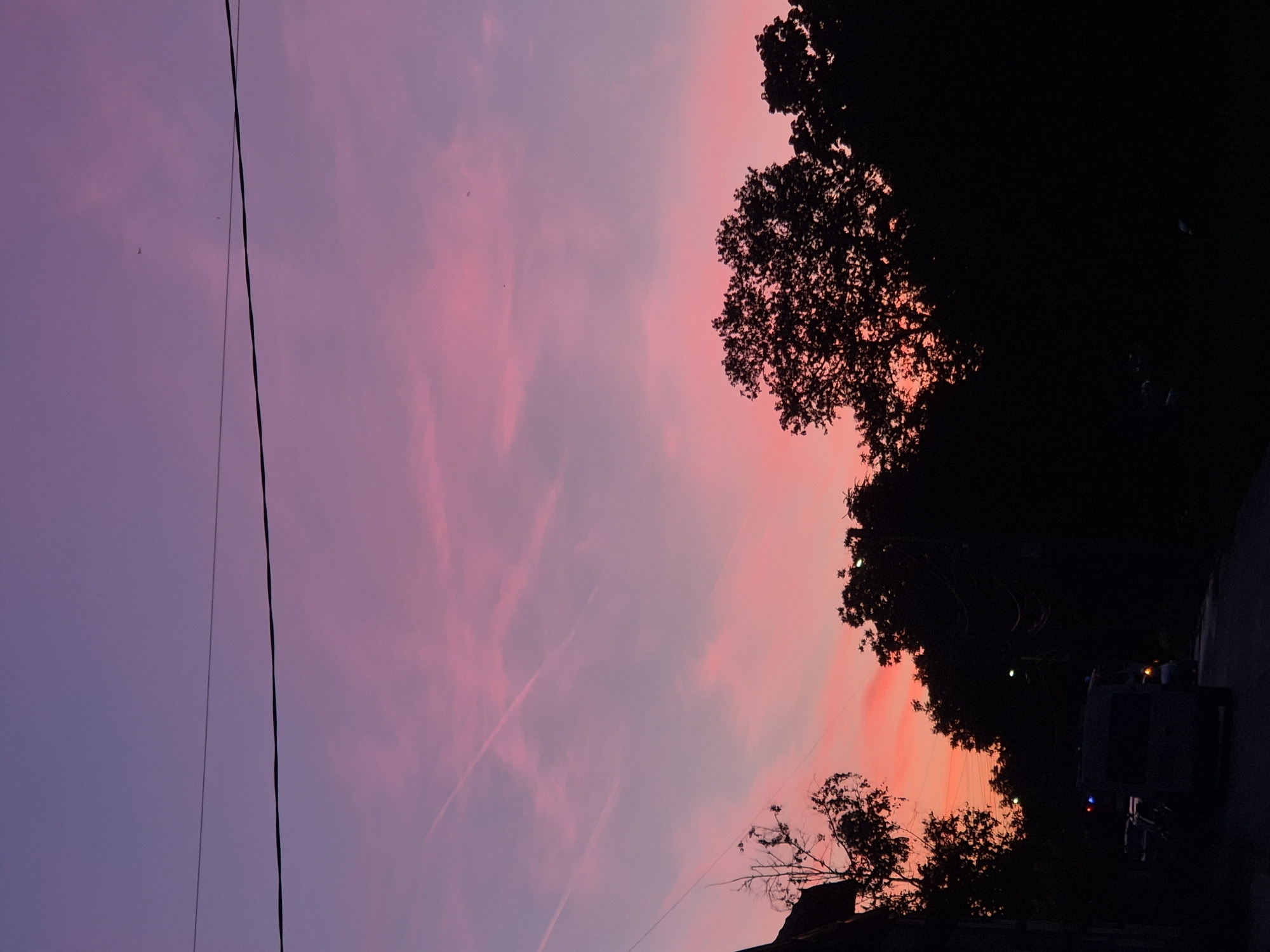 The image depicts a sunset scene in what appears to be the city of Pavlikeni, Bulgaria, though this cannot be definitively confirmed from the image alone. The sky transitions from purple in the upper left to pink and orange hues toward the lower right. A power line runs vertically along the left side of the frame. Silhouetted trees are visible against the colorful sky on the right. There appears to be a structure, possibly a building, partially obscured by trees in the lower center. To the bottom right corner of the image there appears to be a vehicle, maybe a truck with colored lights.